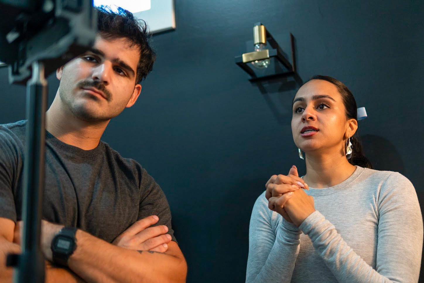 Two people, a man and a woman, are in a room with a dark wall, looking at a camera or screen.