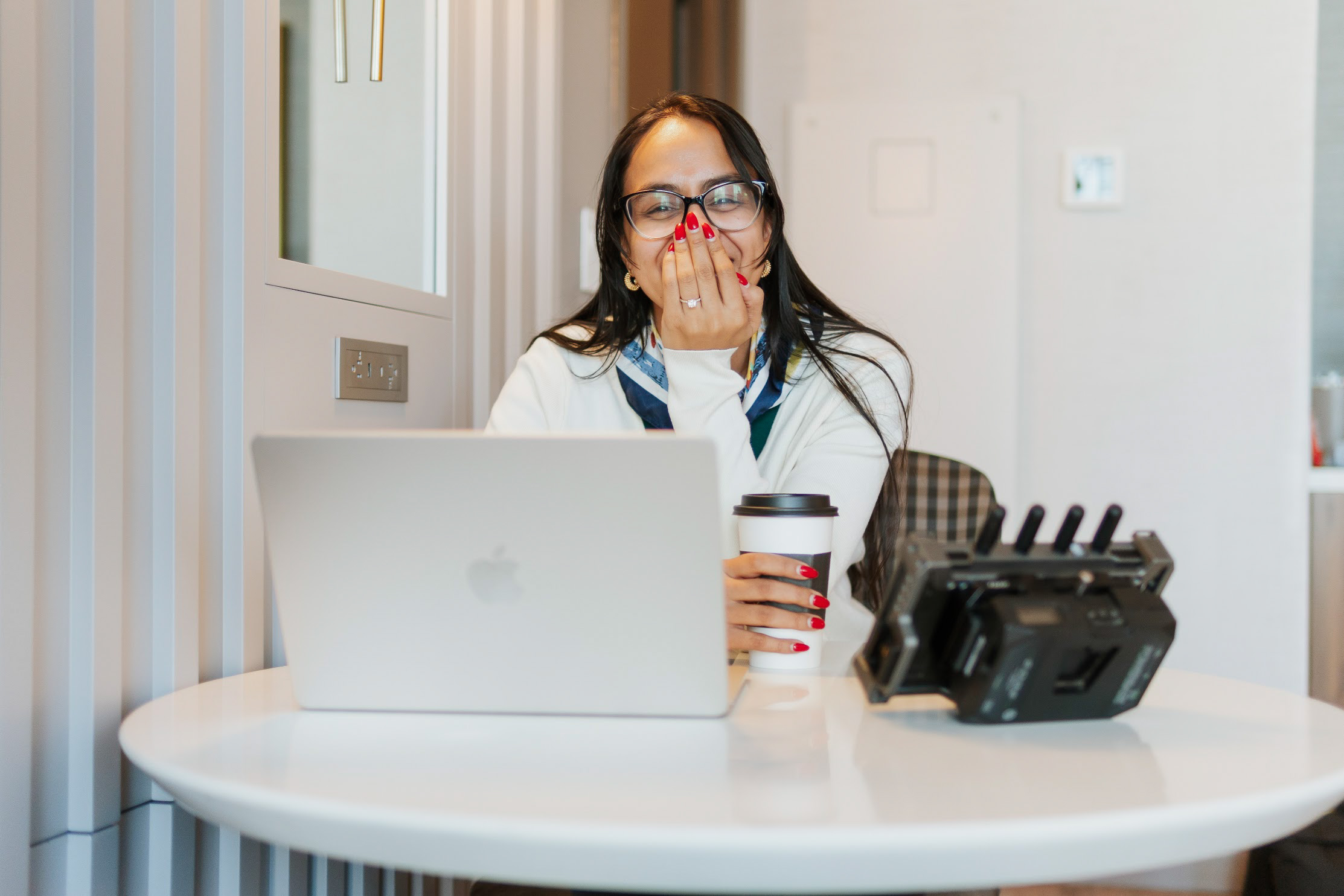 Woman with glasses covering mouth, sitting at a table with a laptop and a coffee cup, holding a phone, in a bright room.