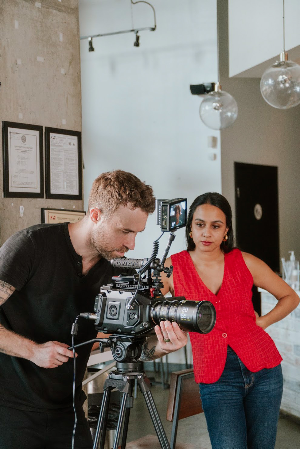 Two people operating a camera in a modern indoor setting with hanging lights and framed documents on the wall.