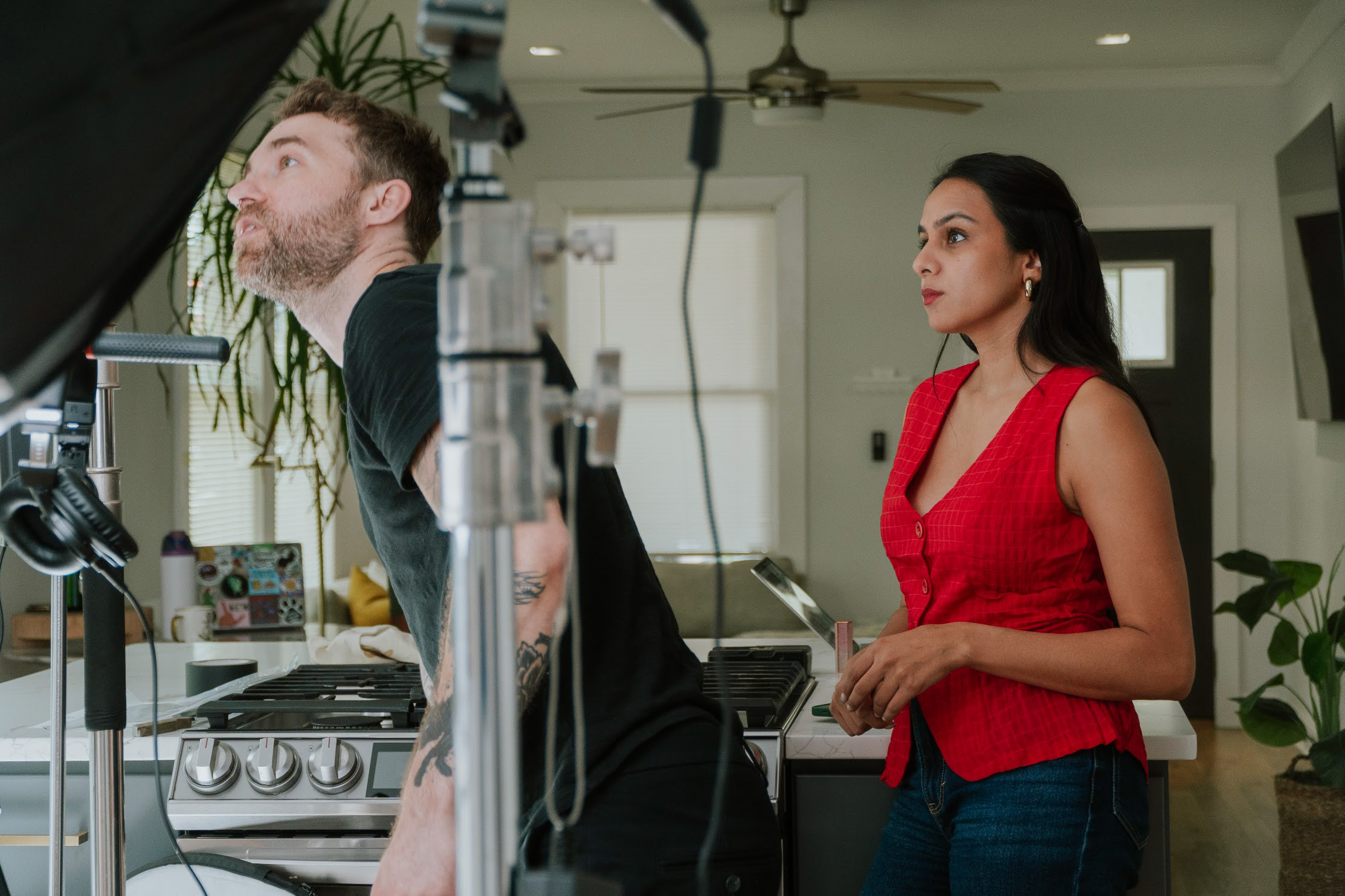 A woman in a red top stands in a kitchen, watching a man working on a film set. Equipment is visible.