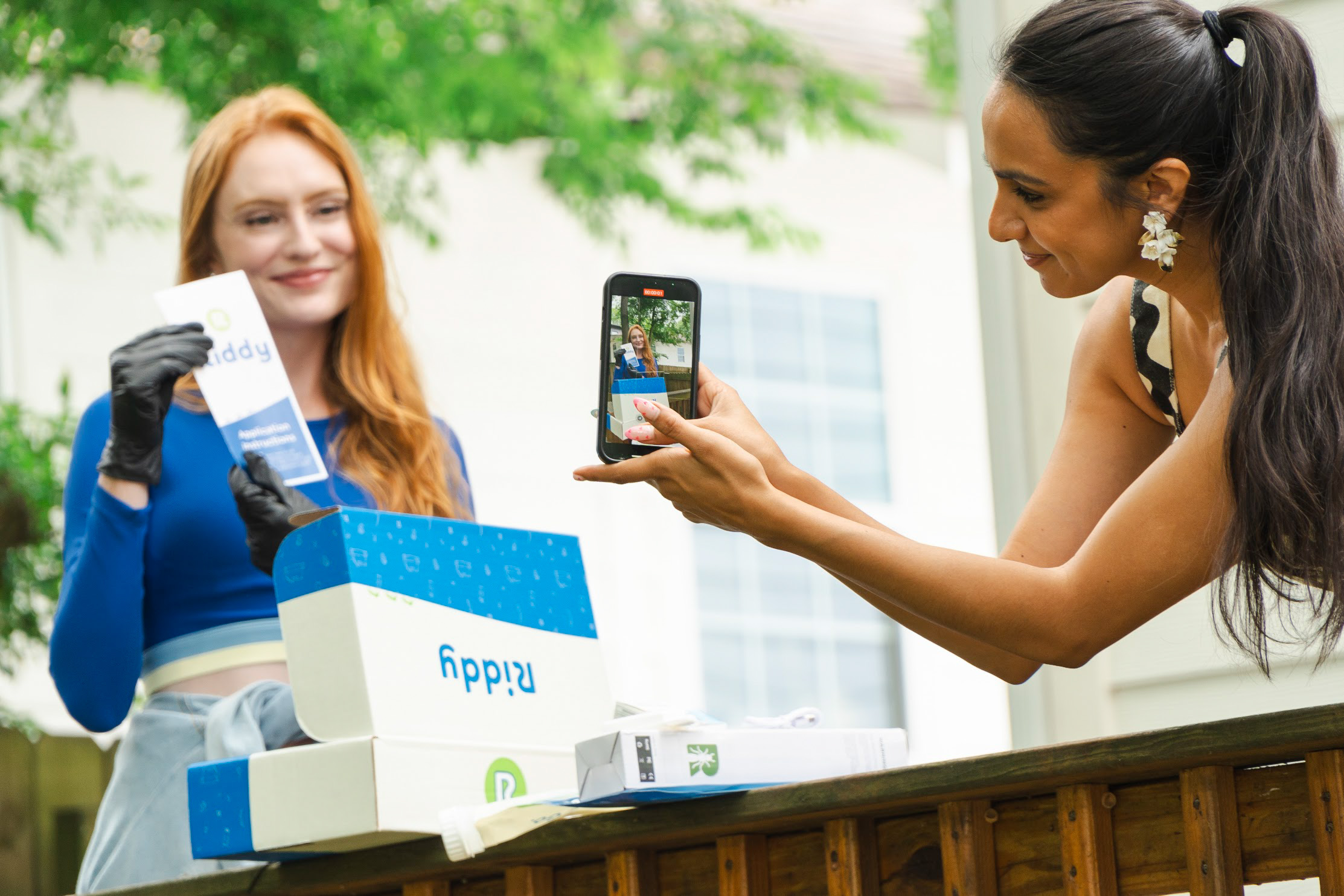 Two women at an outdoor event, one holding a box and the other taking a photo with a smartphone, with trees in the background.