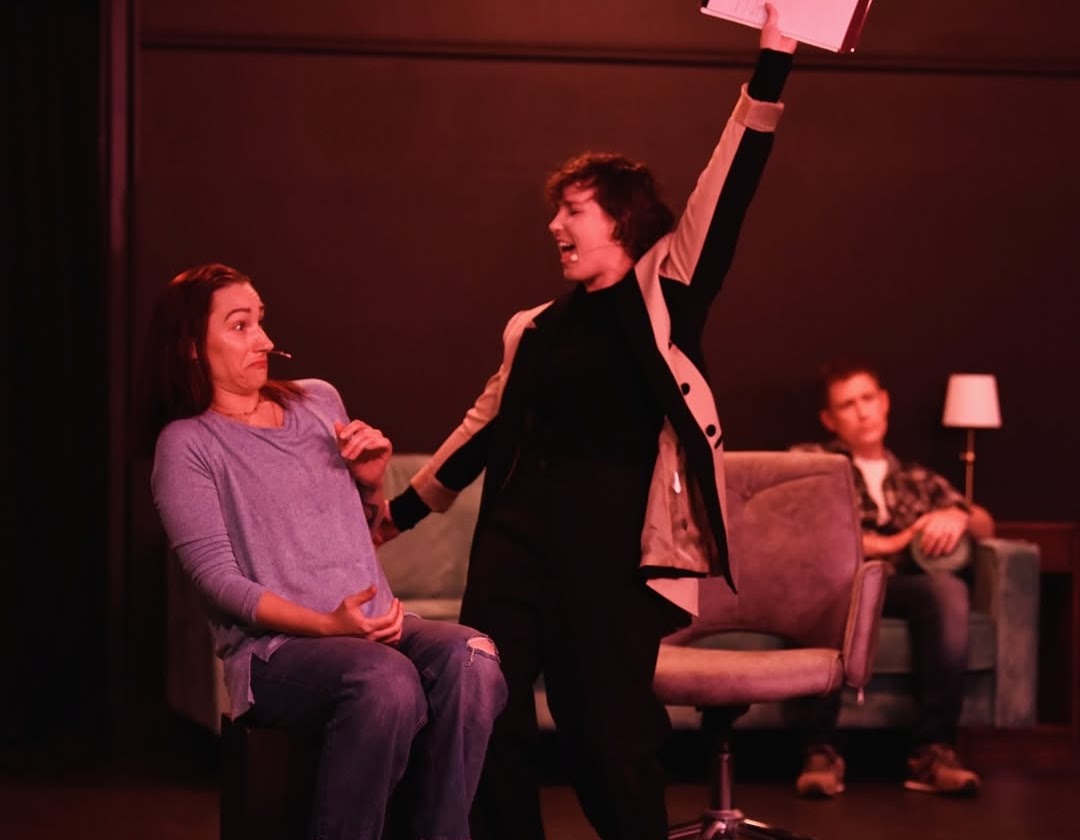 Two women and a man in a room with dark walls; one woman seated, the other woman standing and gesturing excitedly.