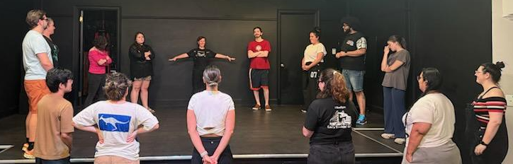 Group of people standing on a stage, some with arms crossed, facing each other in a discussion or rehearsal.