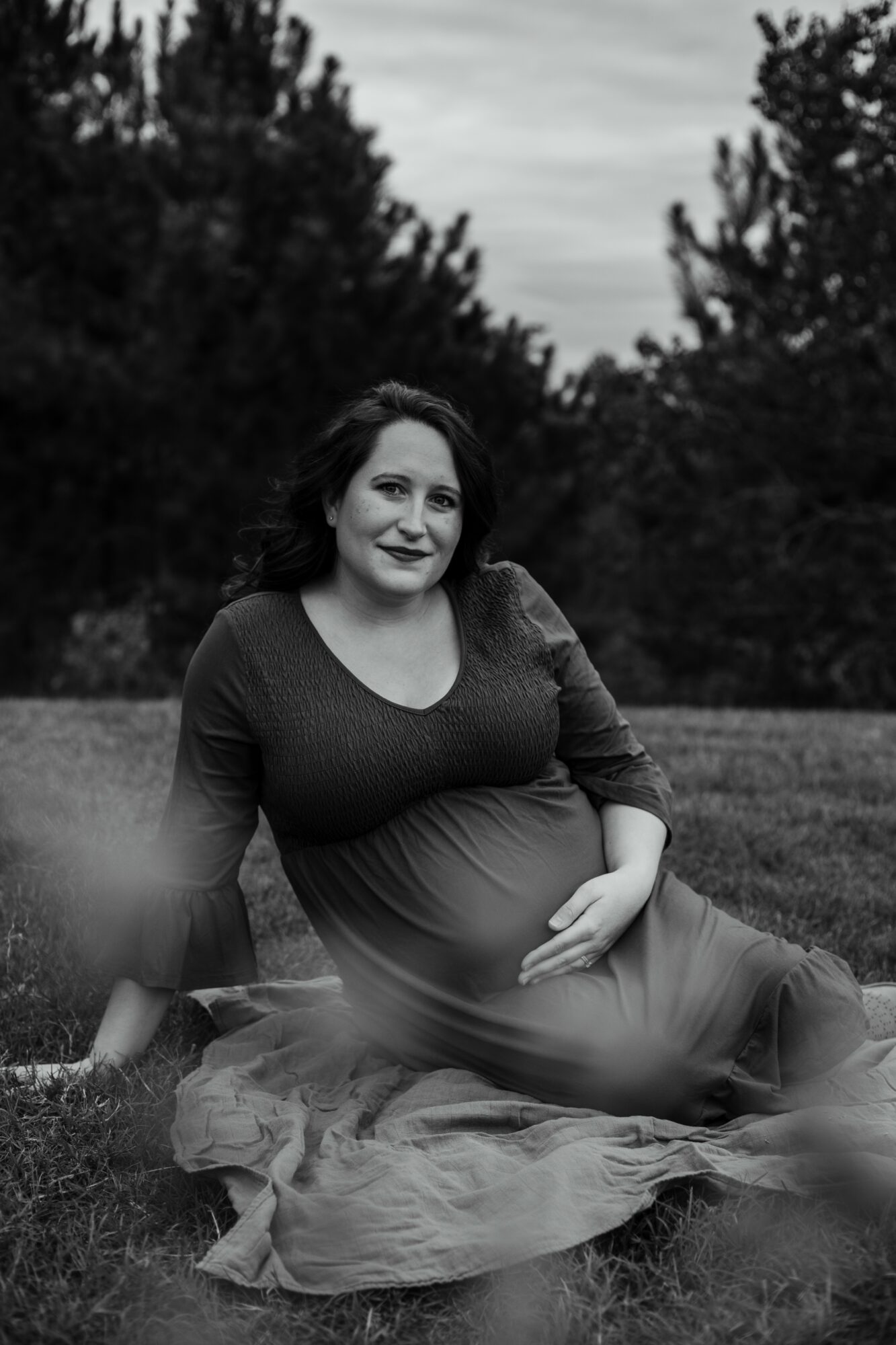 Pregnant woman sitting outdoors on grass, smiling, with trees and sky in background.