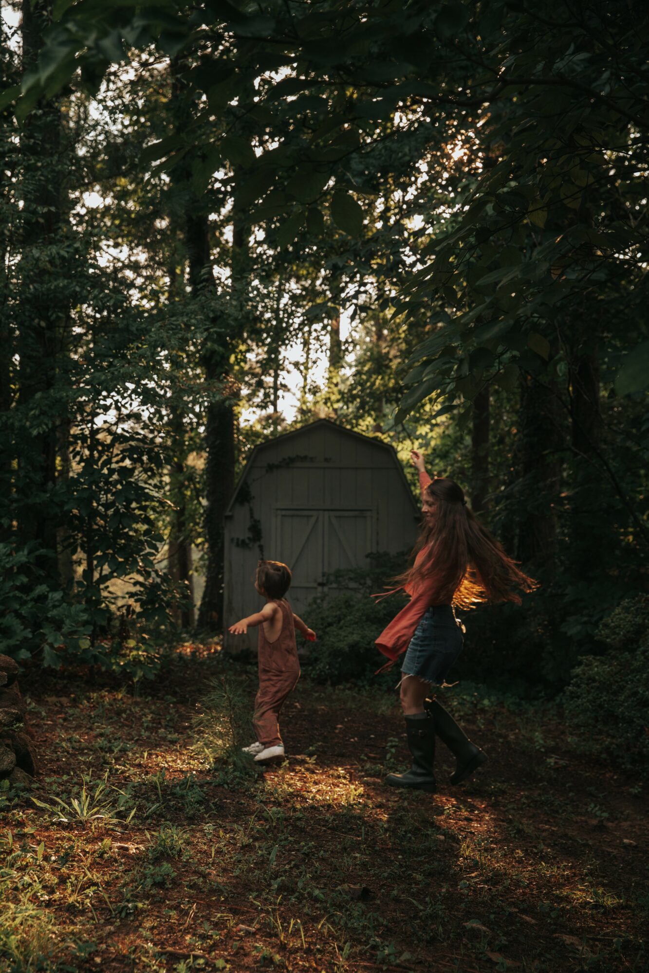 Two children running toward a small shed in a wooded area with sunlight filtering through trees.