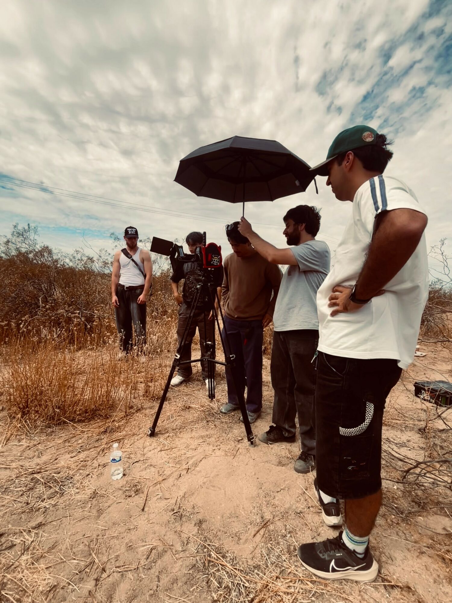 Group of people filming outdoors with camera and umbrella, standing on dry ground under cloudy sky.