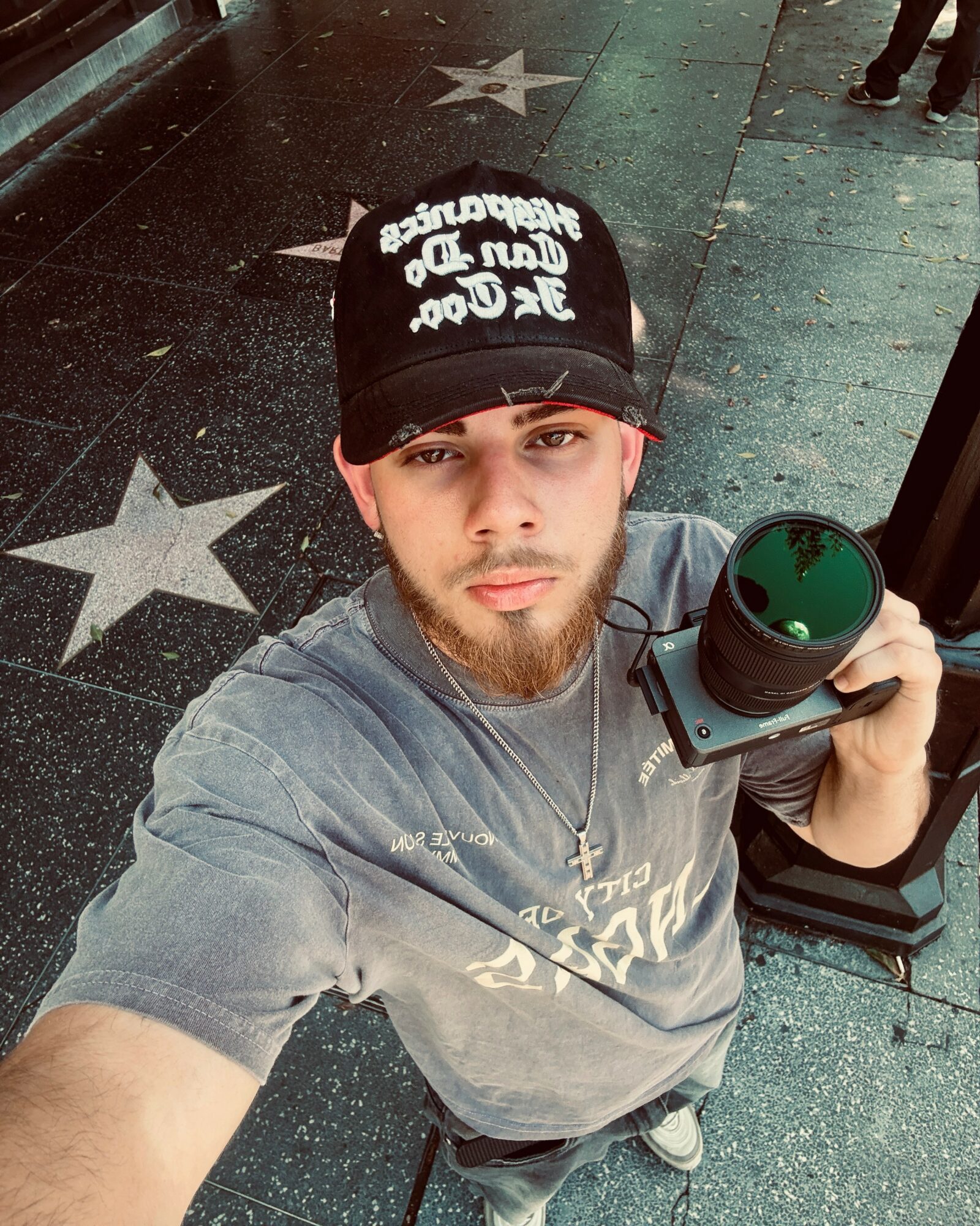 Young man with a beard taking a selfie, holding a camera, wearing a black cap and gray T-shirt, standing on a star-patterned sidewalk.