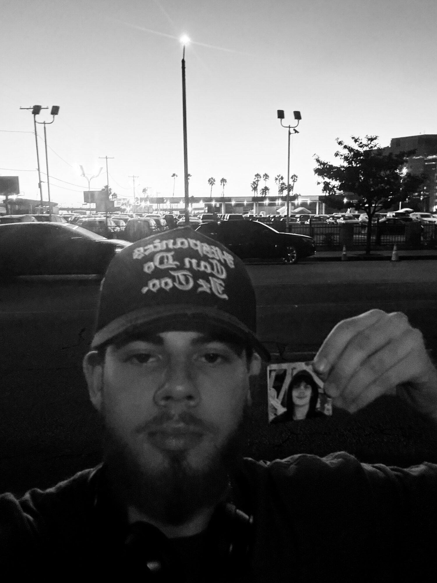 Person holding a photo in a parking lot with streetlights and trees in the background, black and white image.