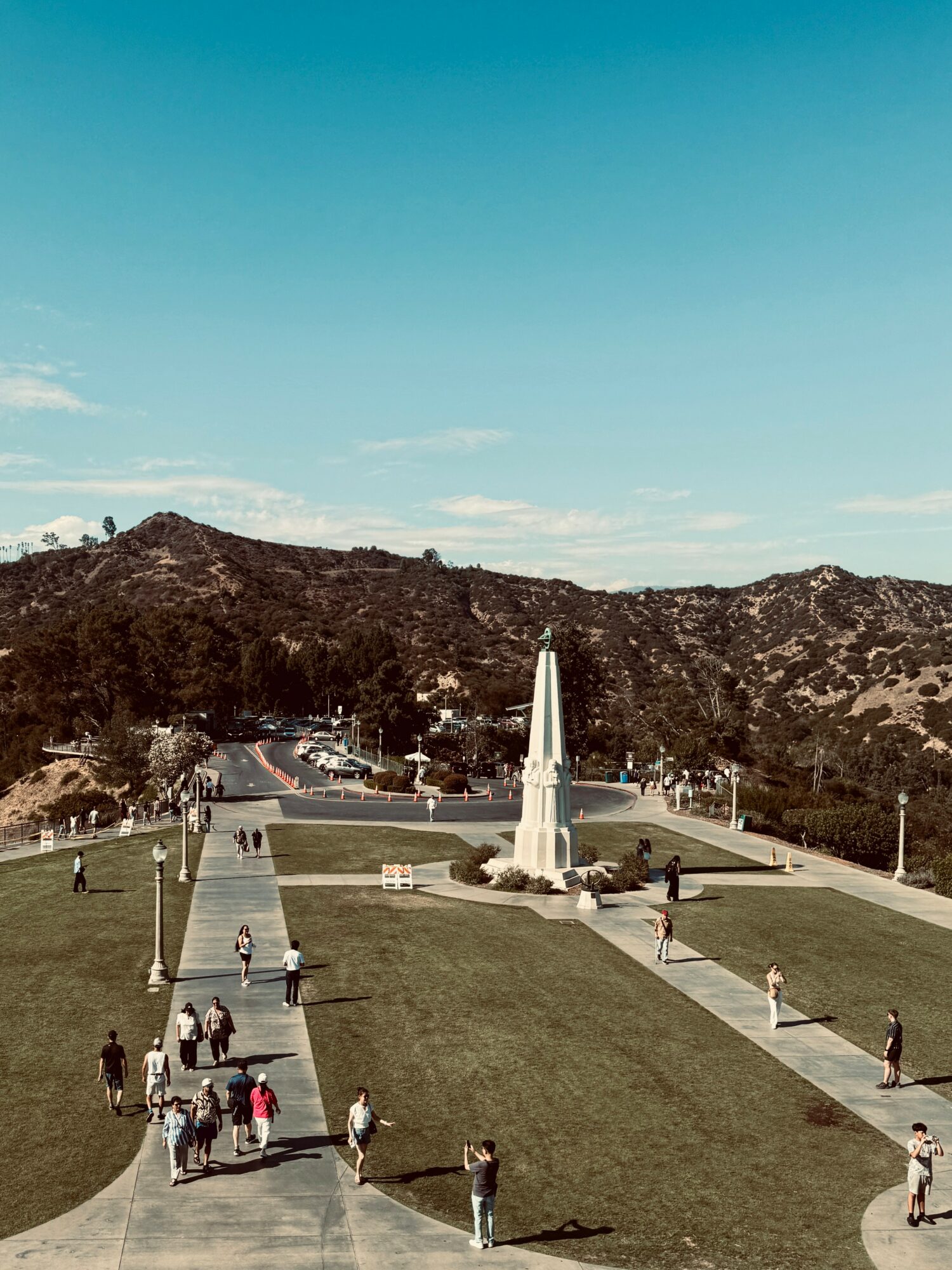 People walk on pathways around a monument on a grassy area with hills and a blue sky in the background.