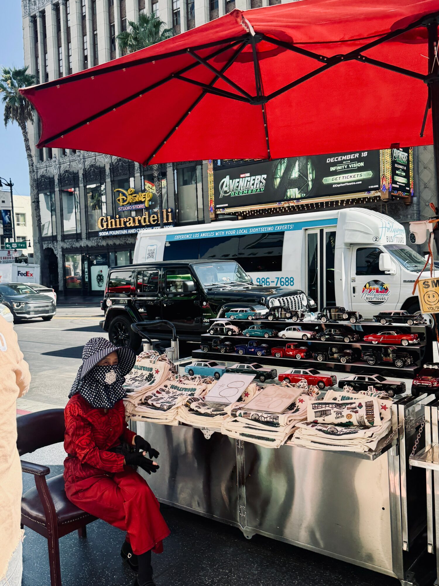 Street vendor selling jewelry under a red umbrella on a busy city street with vehicles and buildings in the background.