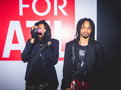 Two people standing in front of a red and white background, one holding a camera, the other with dreadlocks and a black jacket.