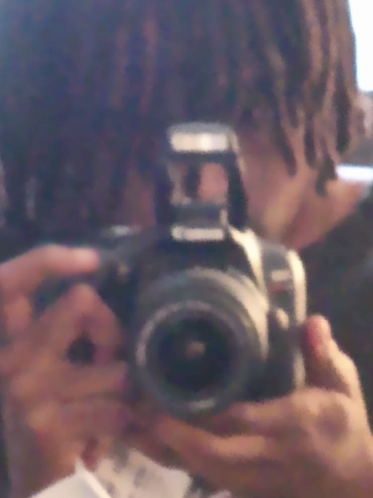 Person with dreadlocks taking a photo with a camera, face partially visible, background blurred.
