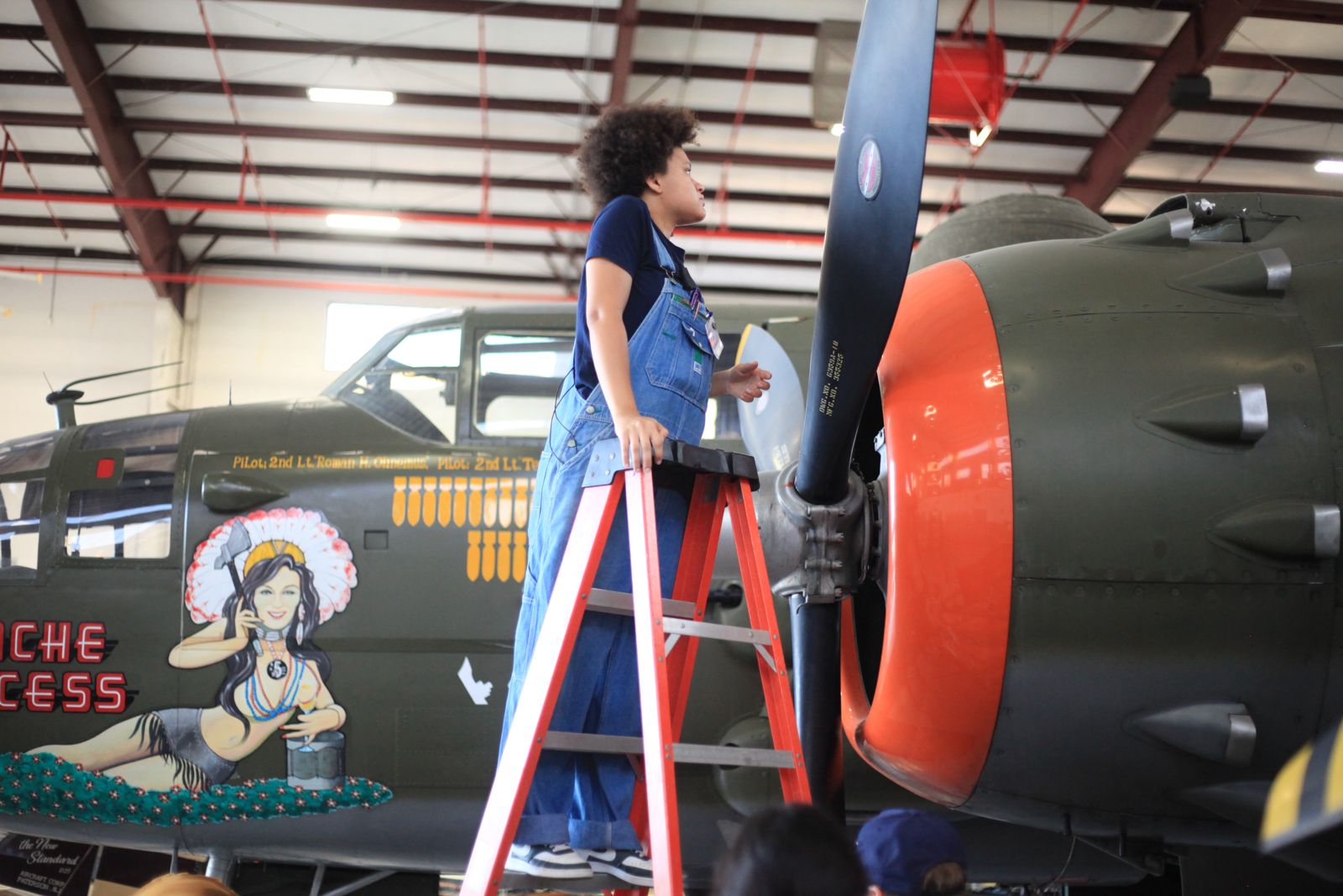 Child standing on a ladder inspecting a vintage military aircraft inside a hangar, with artwork on the plane's side.