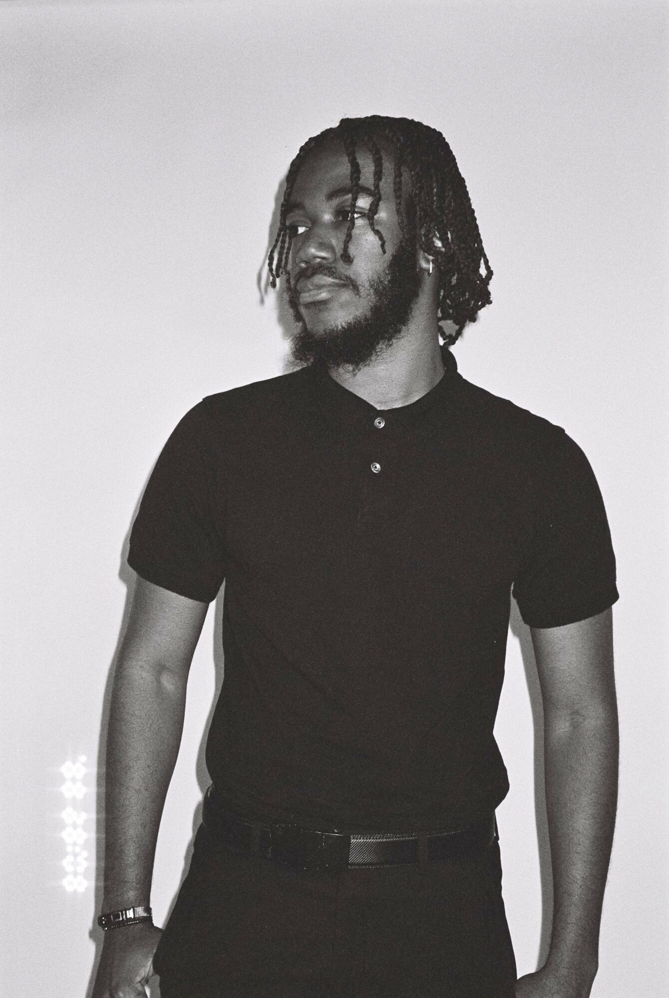 Person with dreadlocks wearing a black shirt and pants, standing against a plain background.