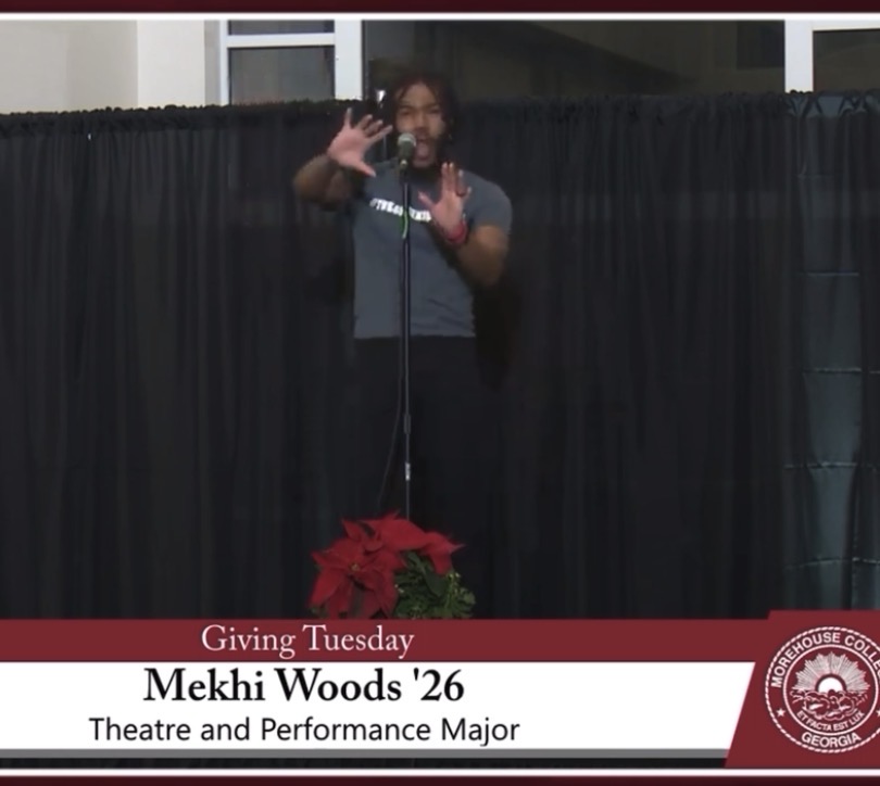 Person standing on stage with a microphone, gesturing with hands, behind a black curtain, with a red poinsettia plant in front.