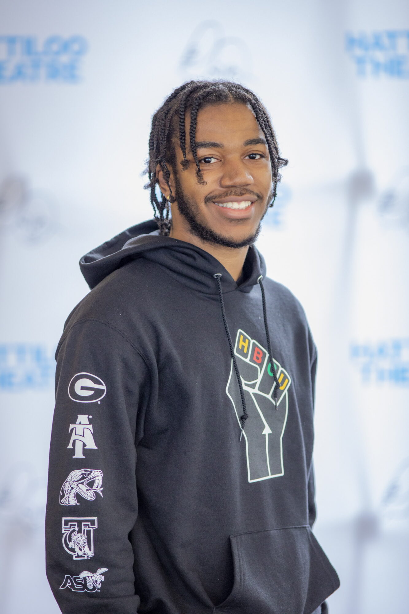 Young man with braided hair smiling, wearing a black hoodie with colorful design, standing in front of a light background.