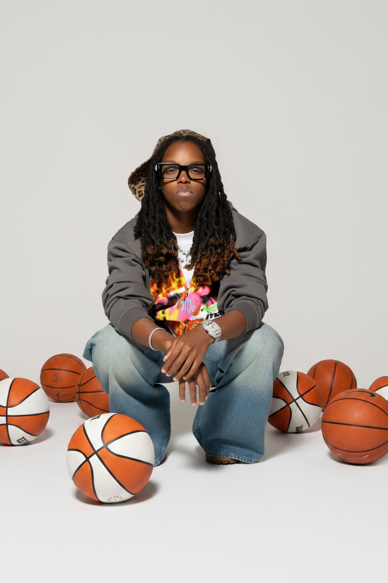 Person with long dreadlocks, glasses, and a colorful shirt, crouching among basketballs on a plain background.