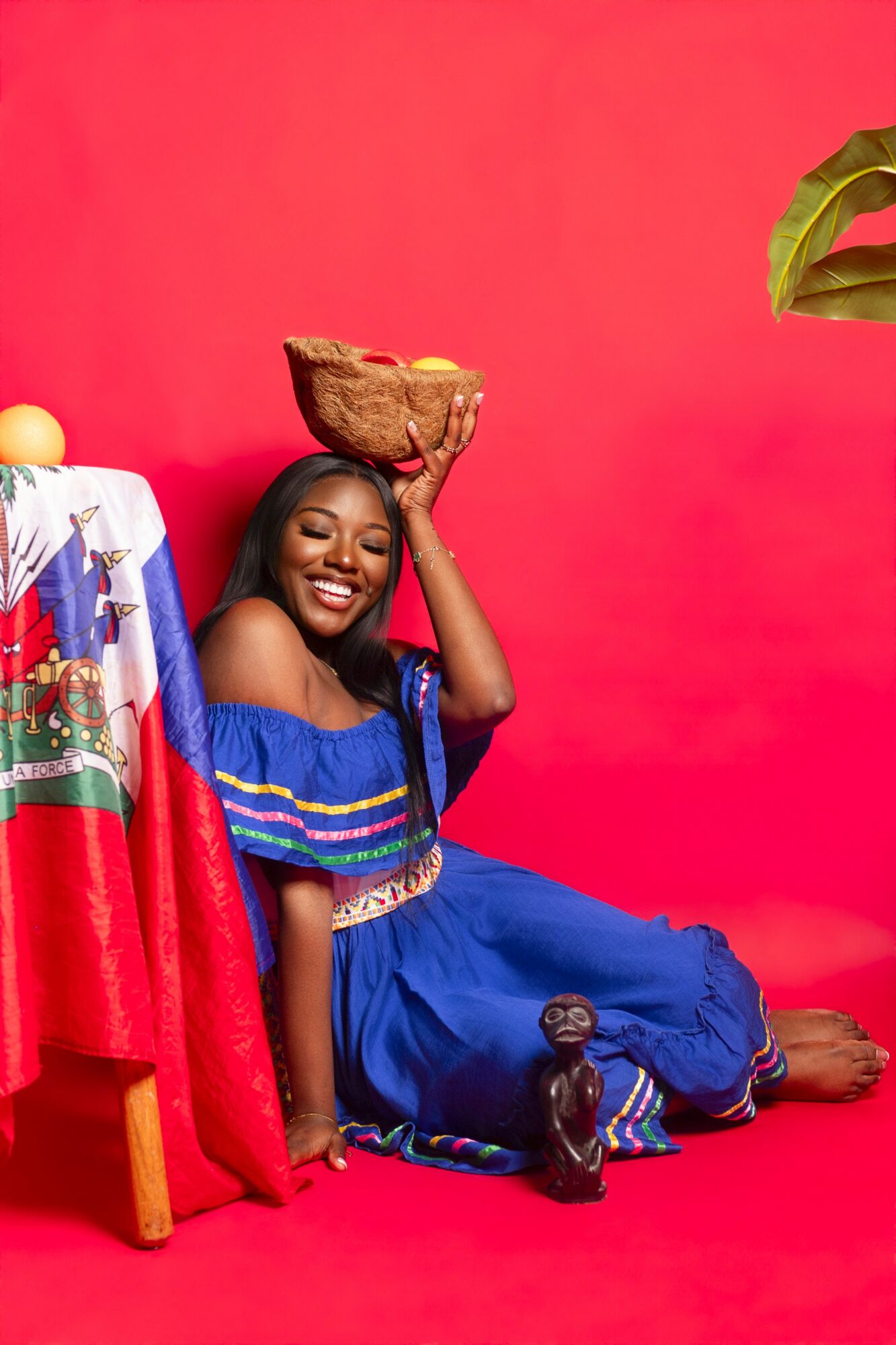 Woman in colorful dress sitting on floor, smiling, holding a basket on her head, with a red background.