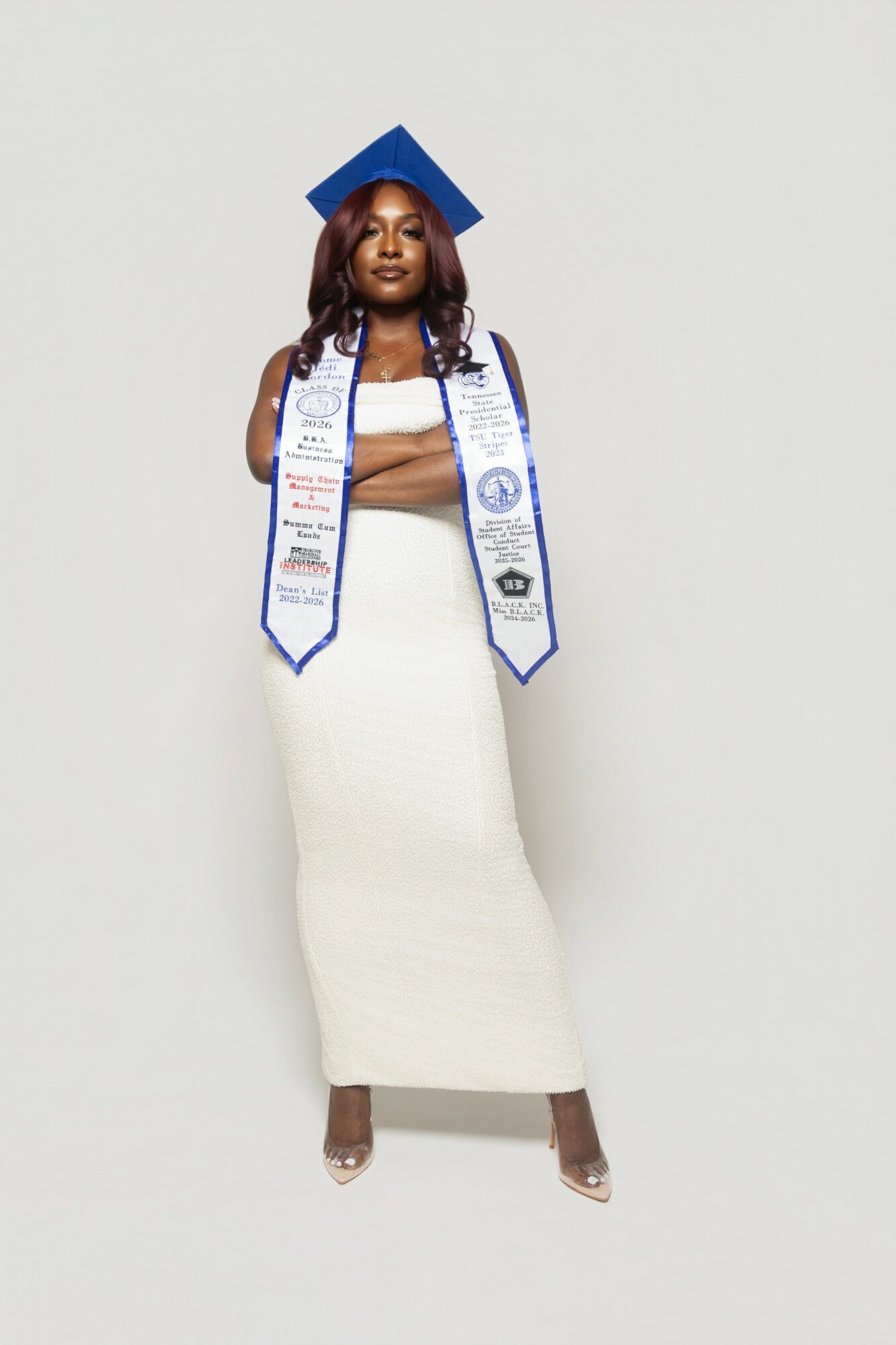 Woman in graduation cap and stole, wearing a white skirt and top, standing against a plain background.