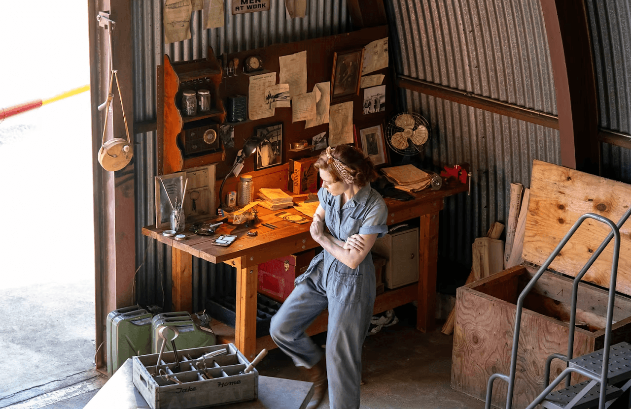 Woman standing in a workshop with a cluttered desk, wooden walls, and various tools and papers, looking down thoughtfully.