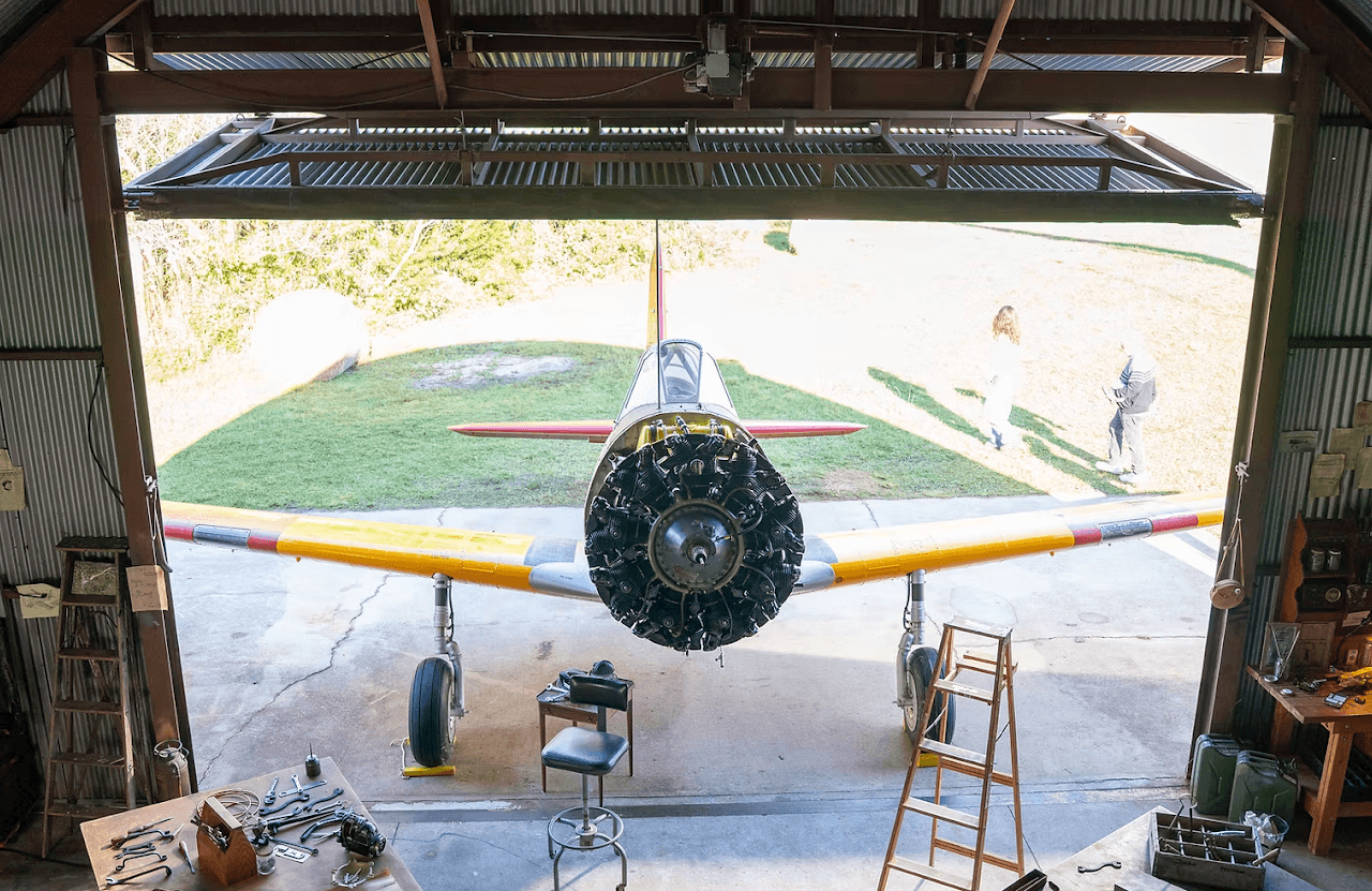 A vintage airplane inside a hangar, viewed from the front with open doors, showing the cockpit and wings, with people outside.