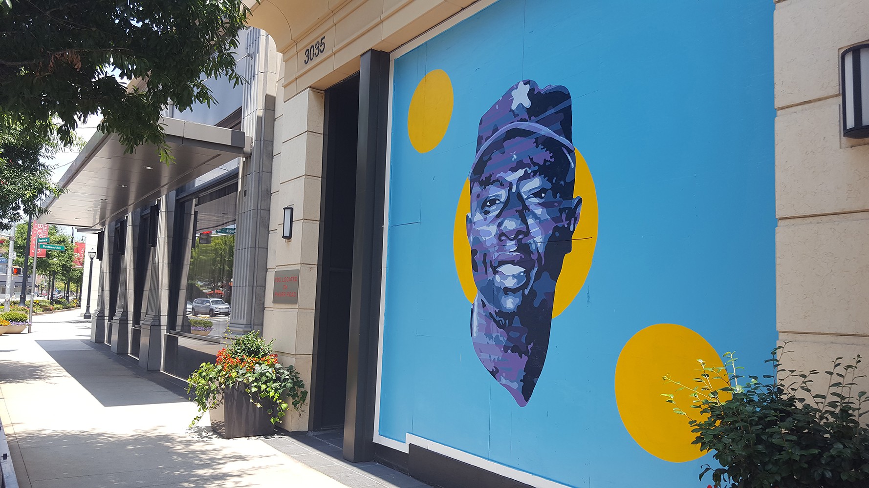 Street view with building featuring a mural of a person's face wearing a hat, surrounded by yellow circles, and trees on sidewalk.