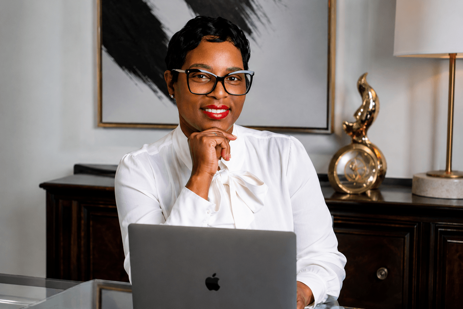Woman with short hair and glasses sitting at desk with laptop, smiling, in a well-lit room with artwork and decor.