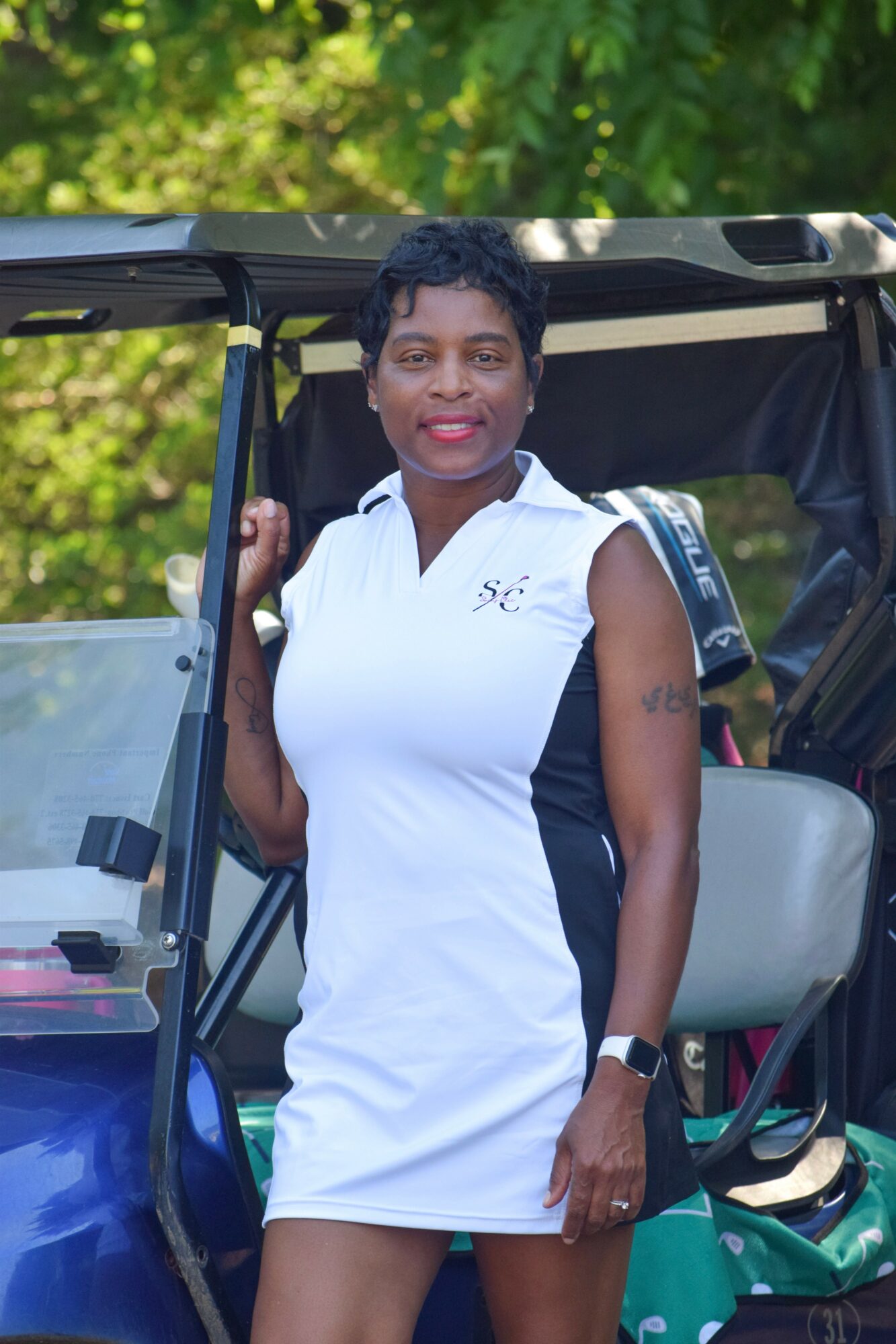 Woman standing outdoors next to a vehicle, wearing a white dress with black accents, smiling, with trees in the background.