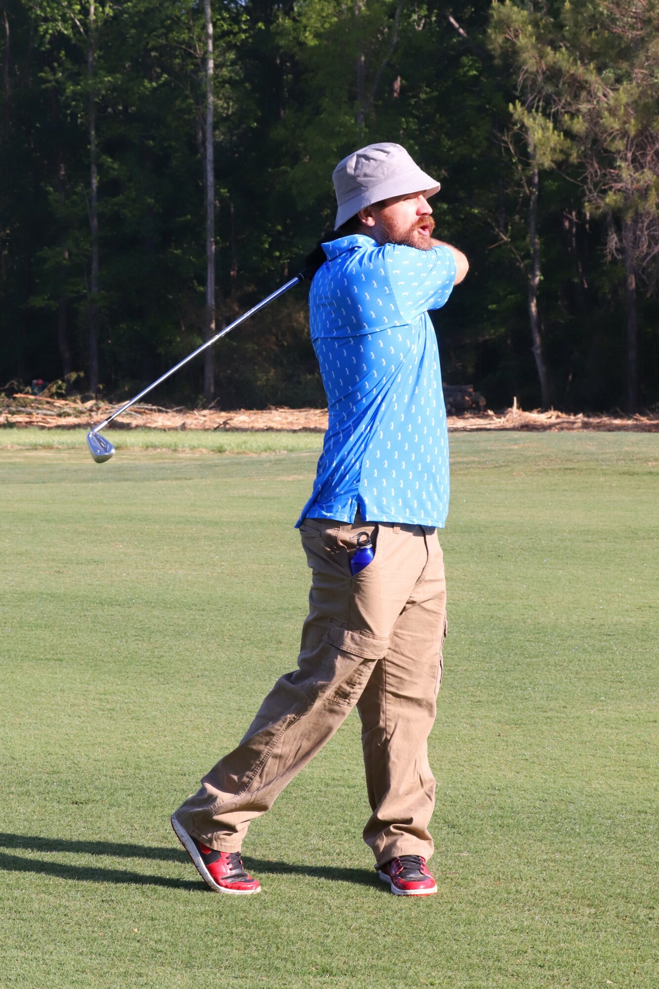 Person walking on a golf course holding a golf club, wearing a hat, blue shirt, and beige pants.
