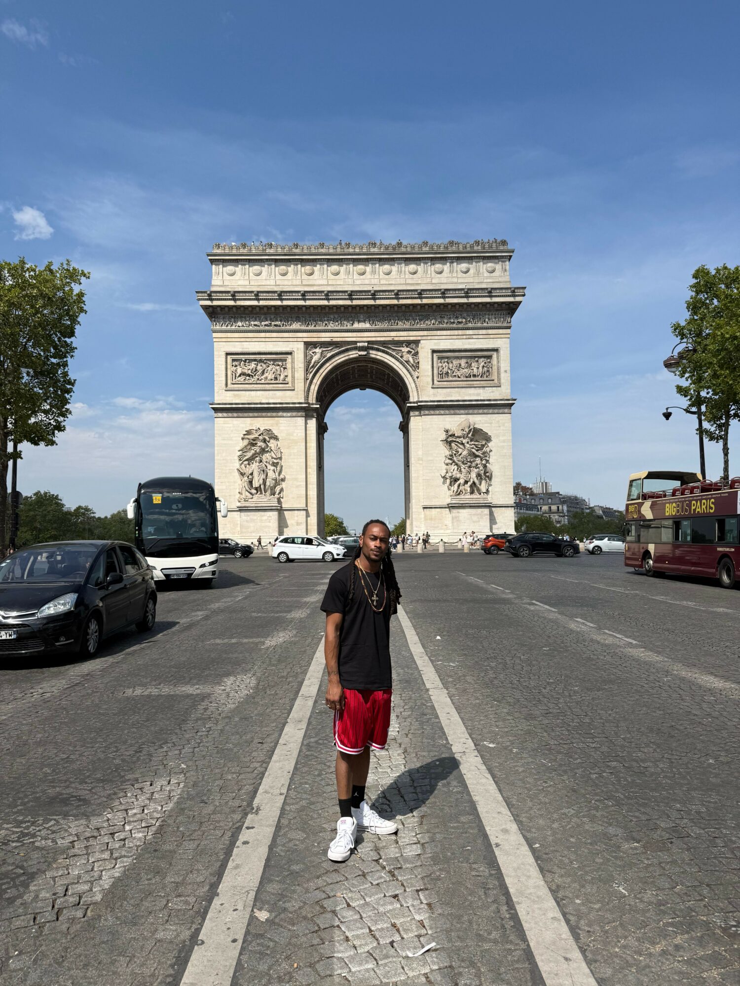 Person walking on street near a large arch monument, with cars parked along the side, trees, and a blue sky.