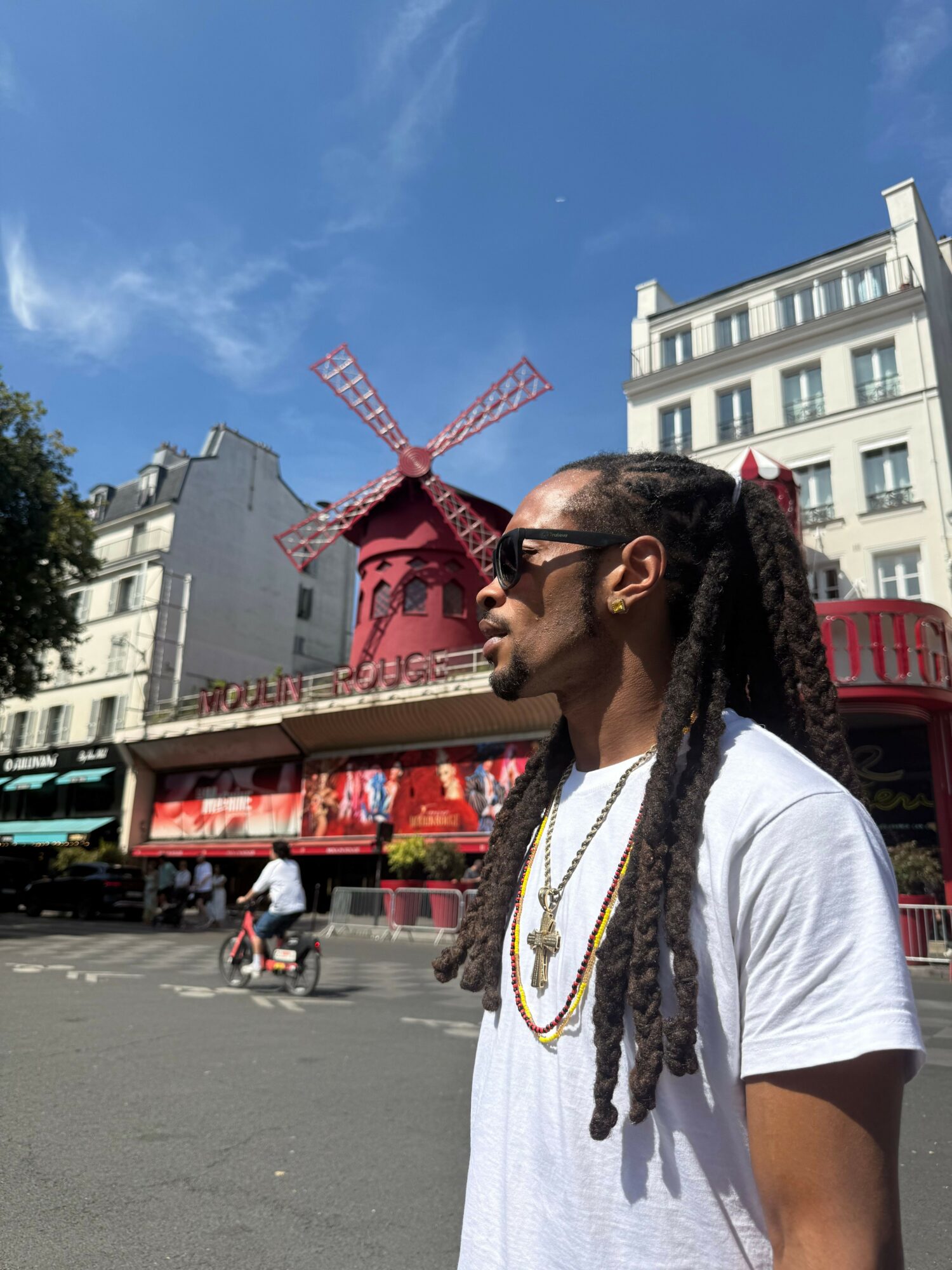 Side view of a woman with long braided hair wearing a white shirt, standing outdoors with buildings and a red windmill in the background.