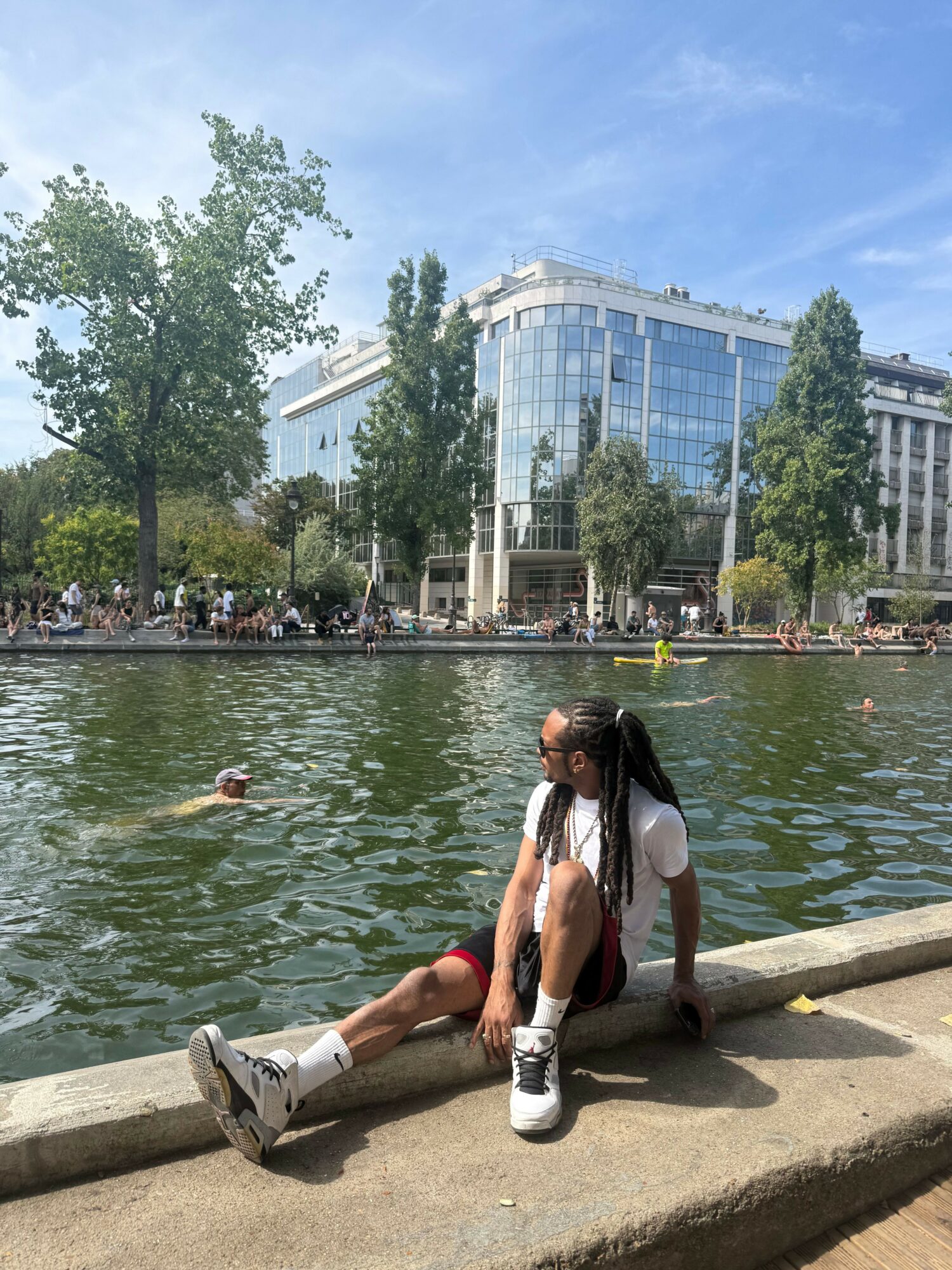 Two women stand on a concrete dock by a body of water, with buildings and trees in the background.