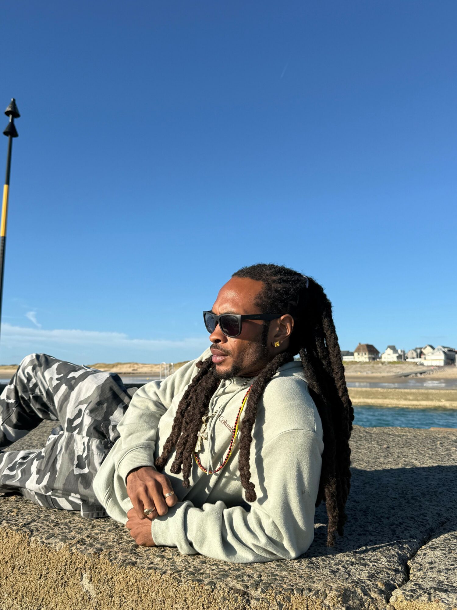 Person with long dreadlocks wearing sunglasses and a light jacket, sitting on rocks near water with a blue sky.
