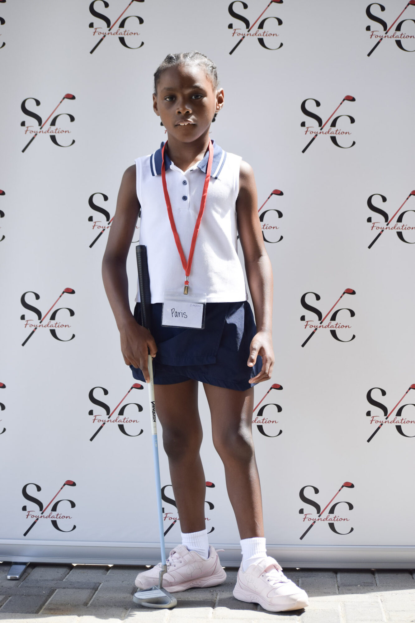 Young girl standing in front of a backdrop with a logo, wearing a white sleeveless top, navy shorts, and white sneakers, with a red lanyard around her neck.