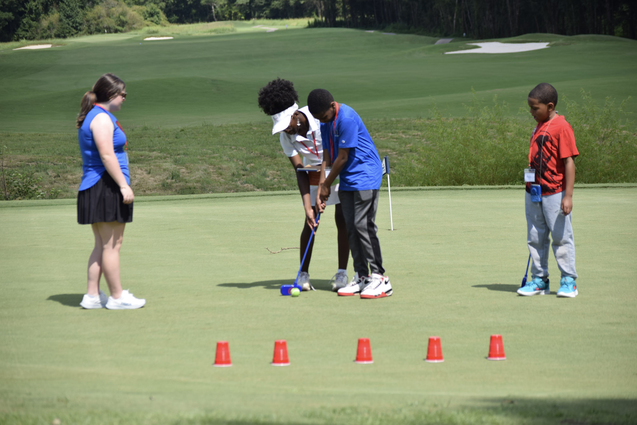 Four children on a golf course practicing putting, with five orange cones in front of them, green grass, and trees in the background.