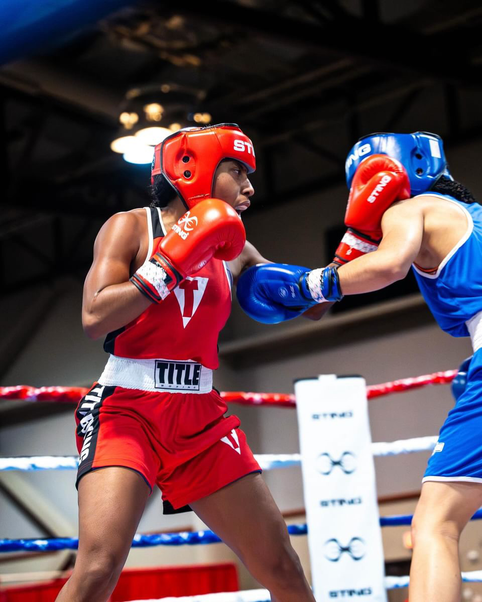 Two boxers in red and blue gear sparring in a boxing ring, with a dark background and overhead lights.