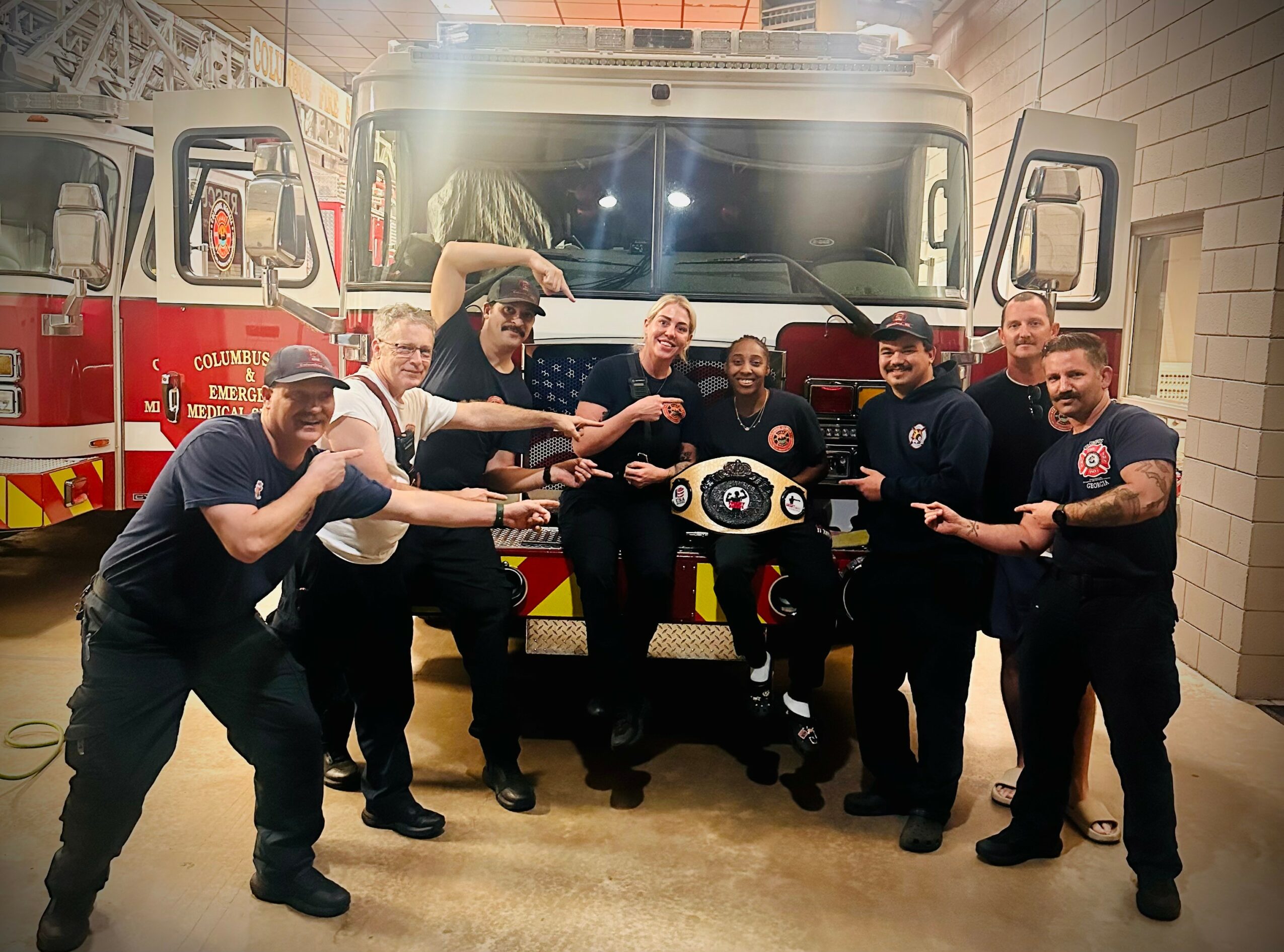 Group of seven firefighters in uniform posing in front of a fire truck inside a garage, smiling and pointing at a small vehicle model.
