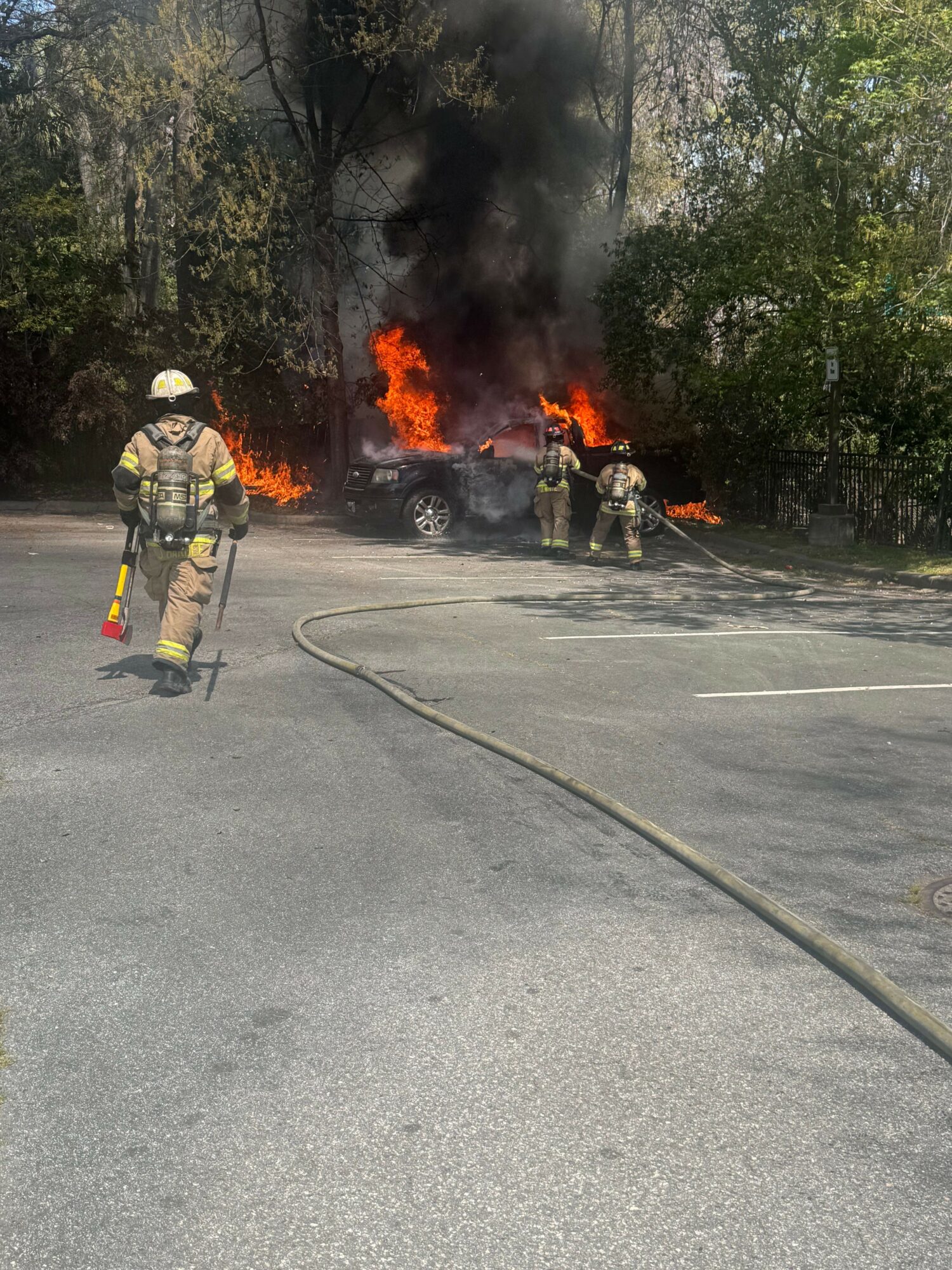 Firefighter in protective gear stands near a burning vehicle with flames and smoke, holding a hose in a parking lot.