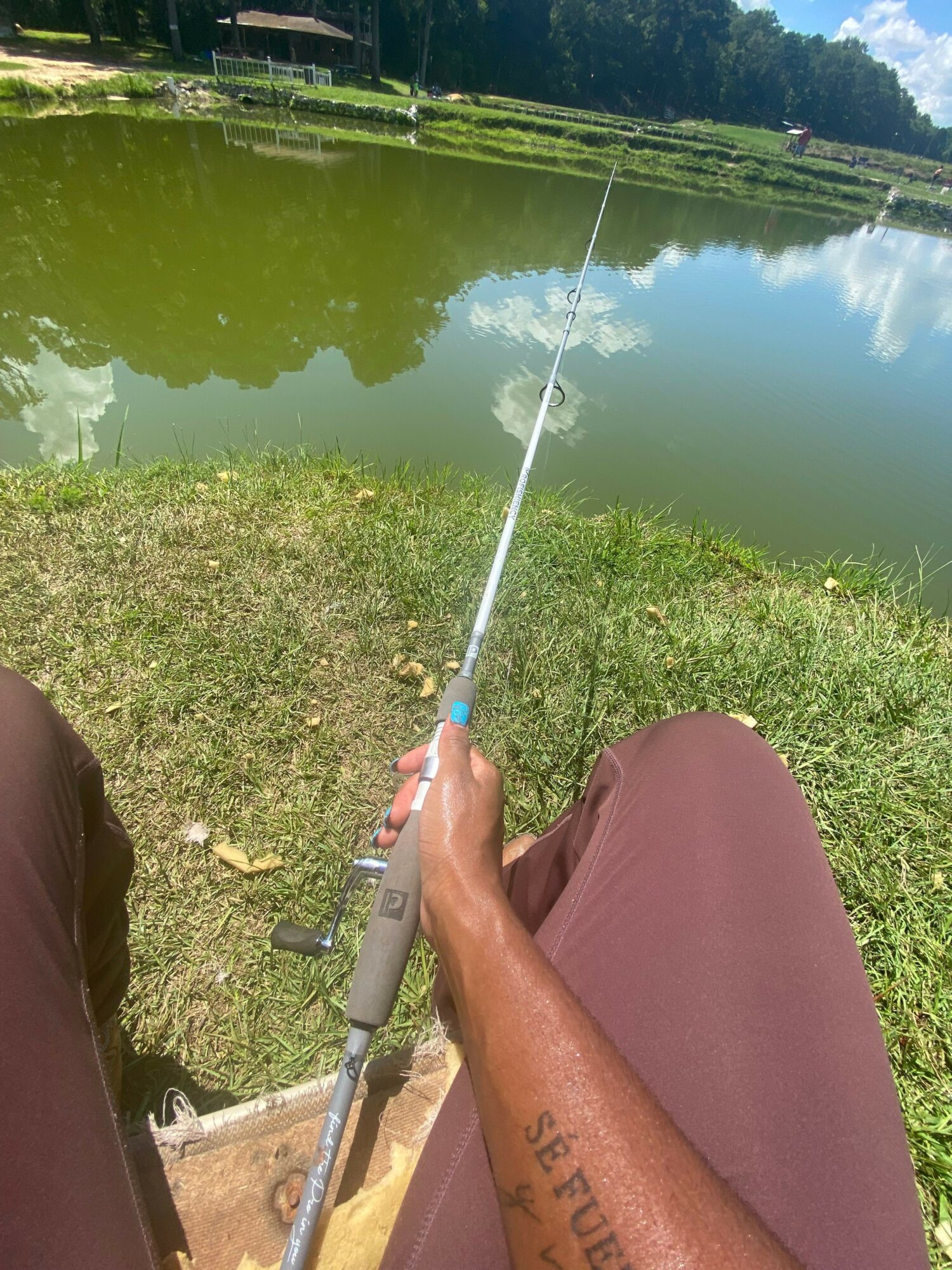 Person holding a fishing rod near a body of water with green grass and trees, cloudy sky reflected in the water.