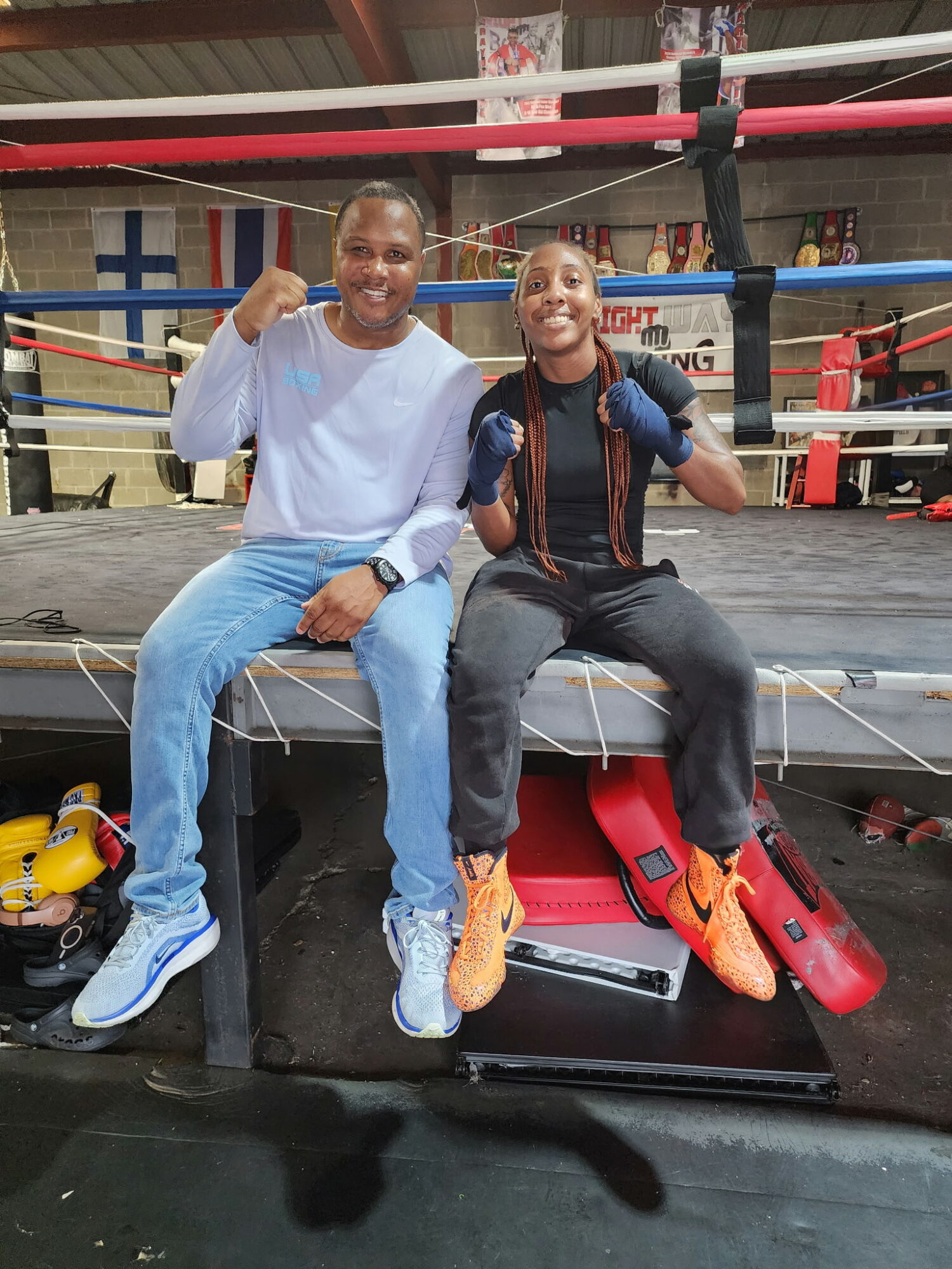 Two people sitting on the edge of a boxing ring, smiling and raising fists, with boxing gloves on the floor nearby.