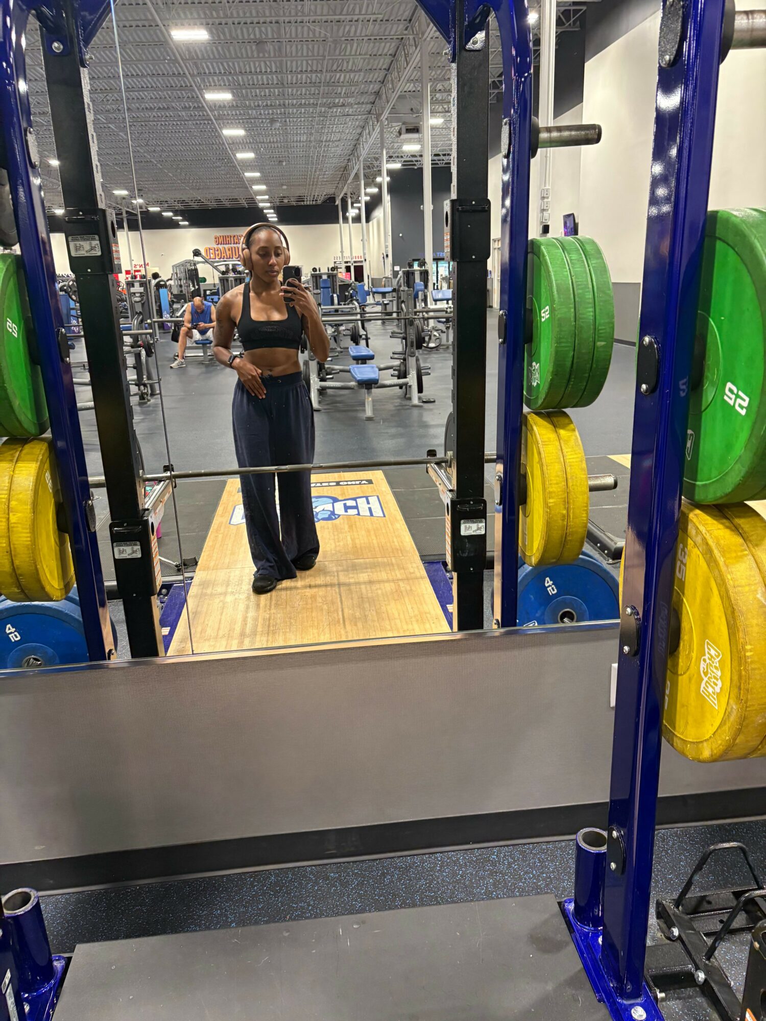 Person taking a mirror selfie in a gym, with weight plates on racks and gym equipment visible.