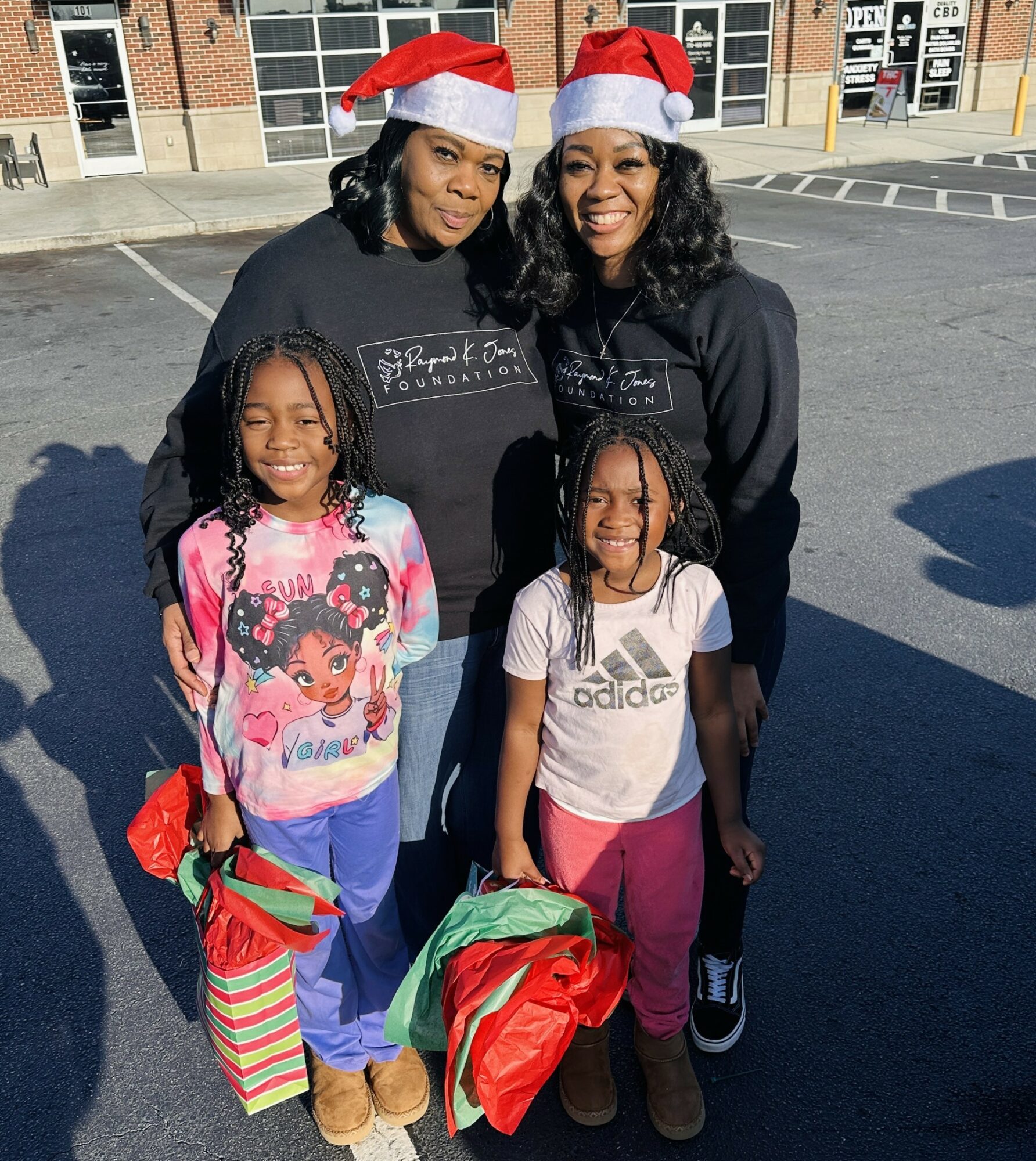 Two women and two children wearing Santa hats stand outdoors in a parking lot, smiling, with buildings in the background.