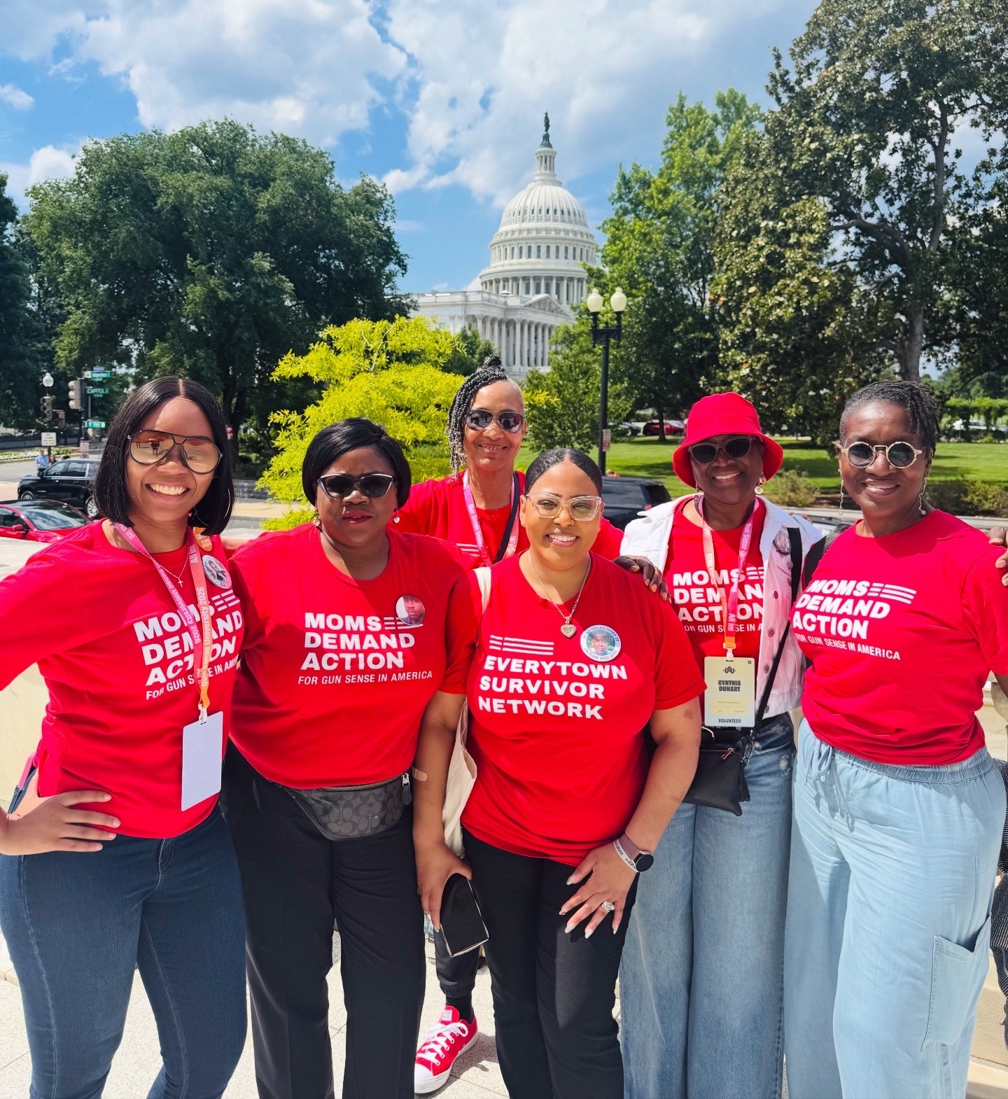 Six women wearing red shirts standing outdoors in front of the U.S. Capitol building, smiling, with trees and blue sky behind them.