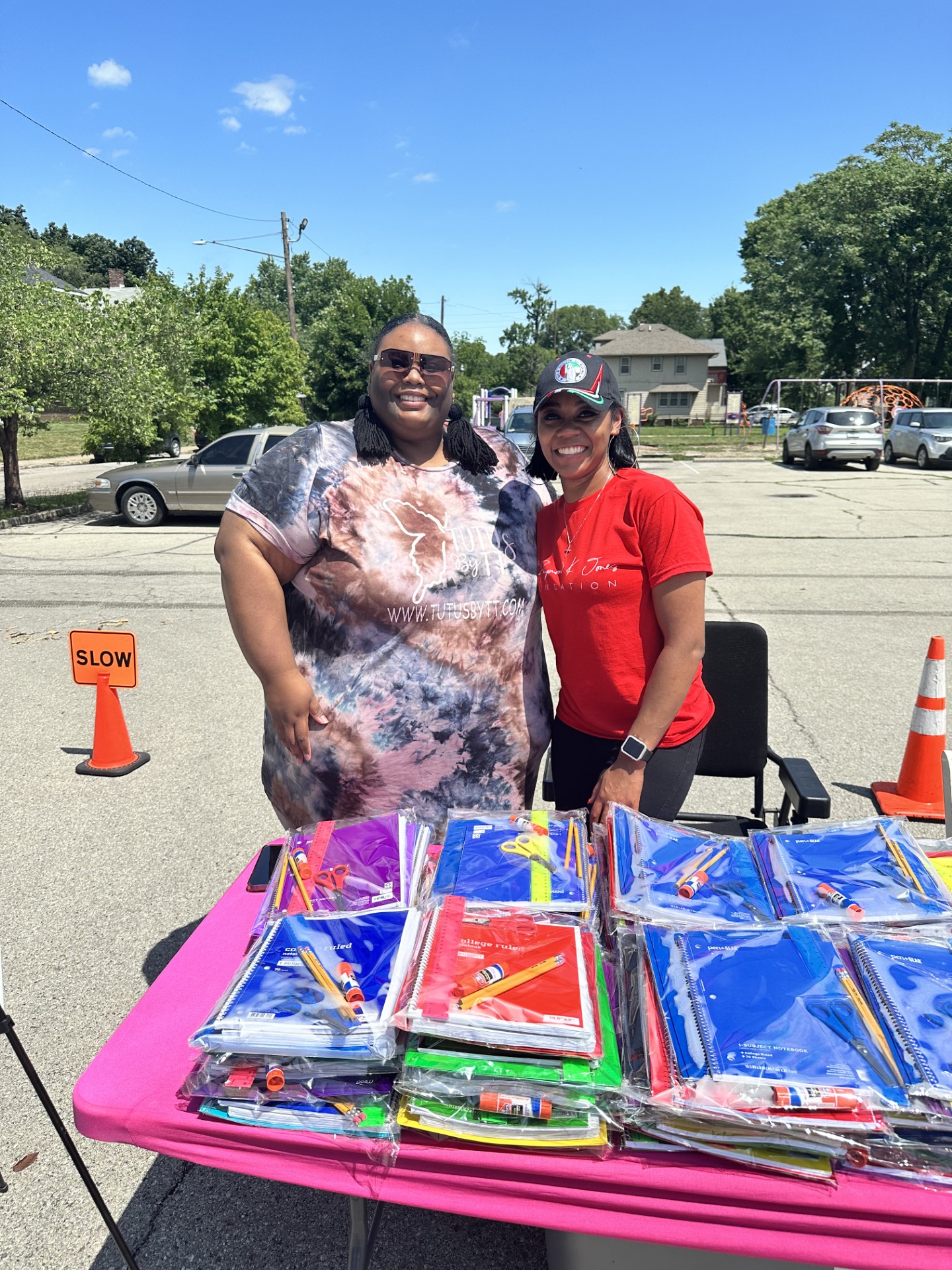 Two women standing behind a pink table with colorful packaged items outdoors on a sunny day, with cars and trees in background.