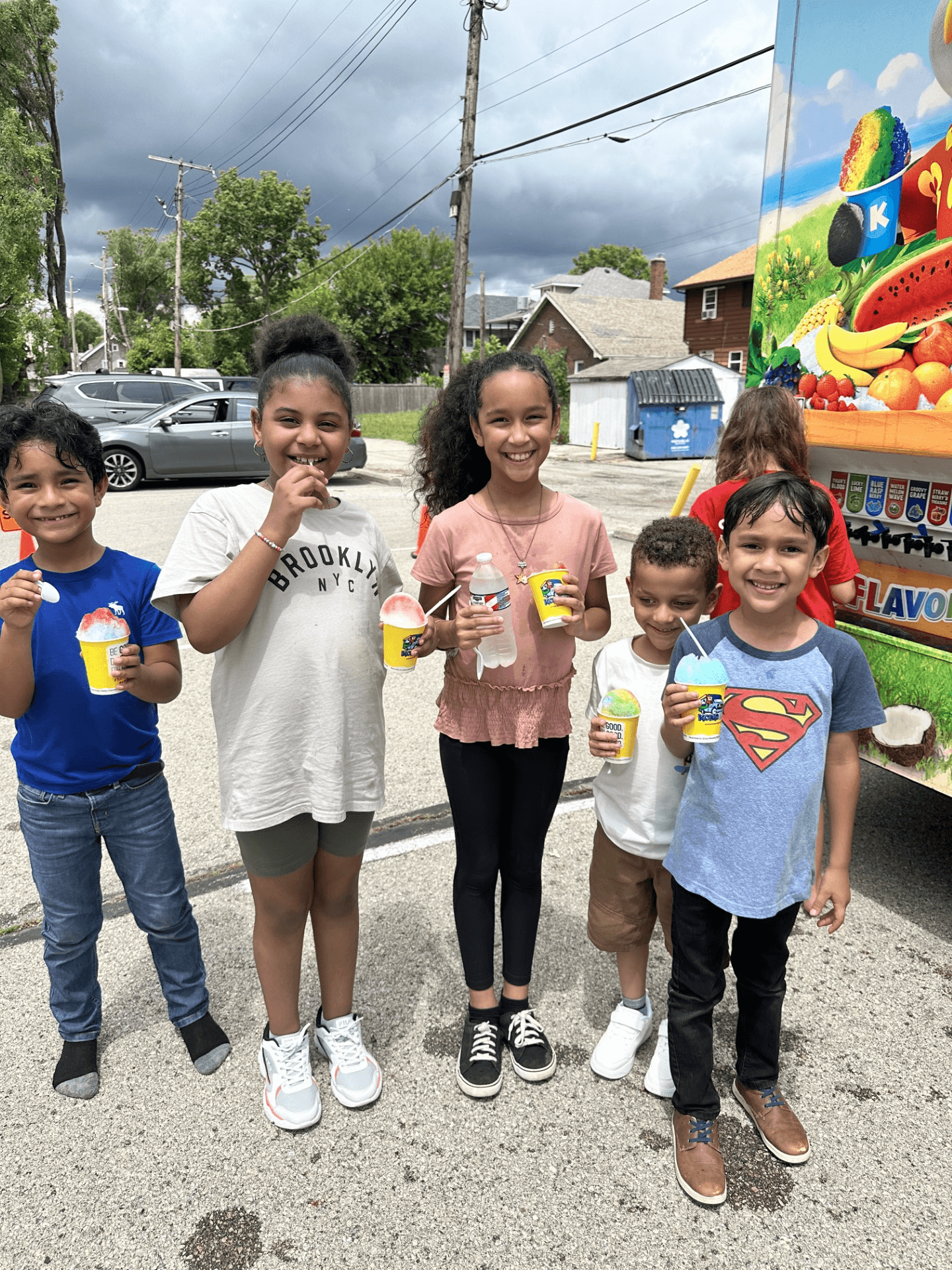 Group of five children standing outdoors, holding ice cream cones, smiling, with a colorful vending machine behind them.