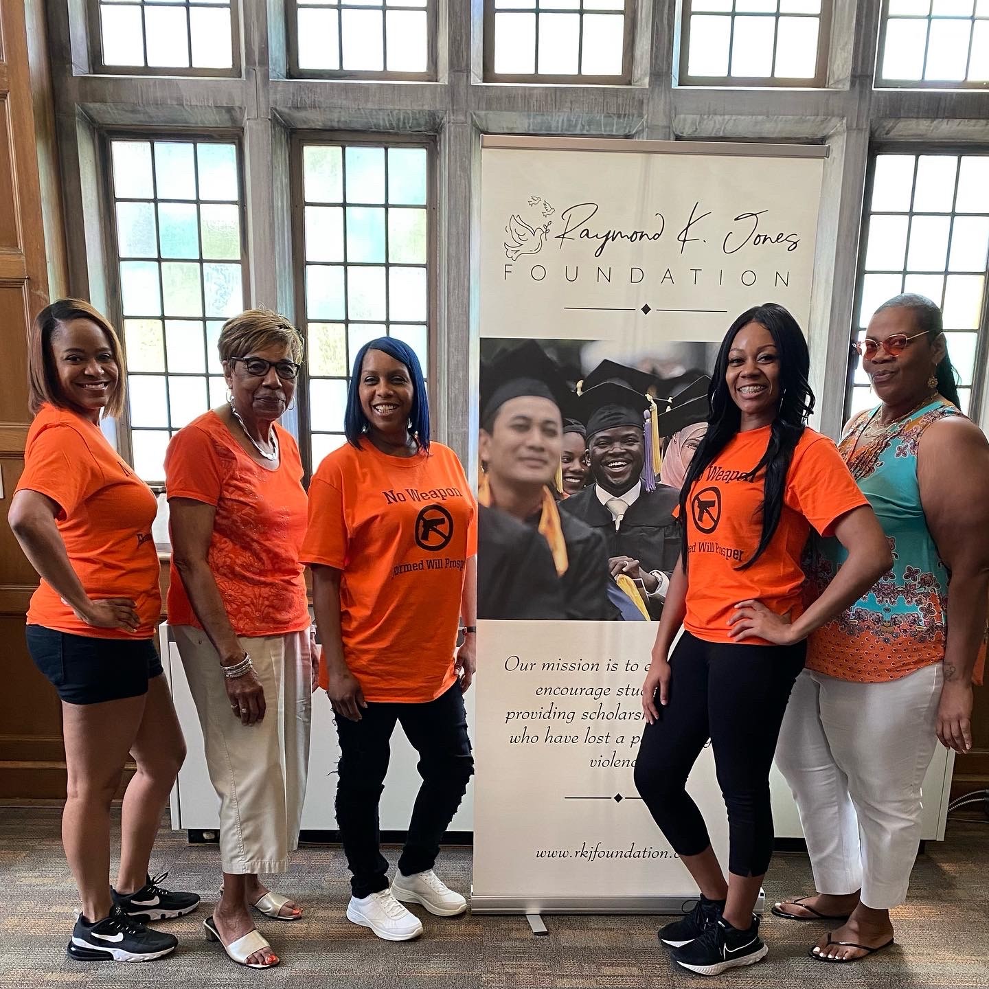 Five women stand in front of a large window and a banner, smiling for the photo, indoors.