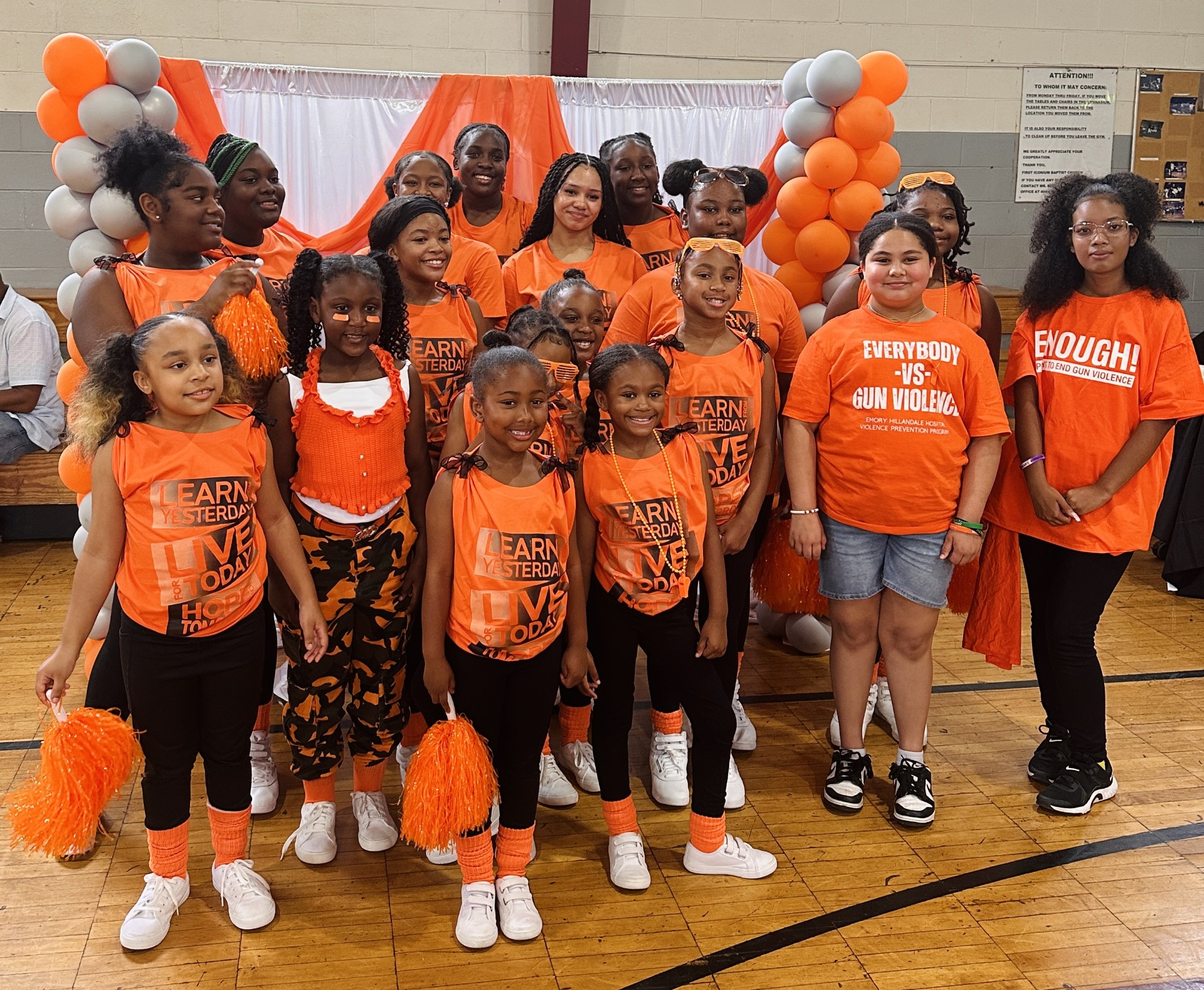 Group of children and adults in orange shirts and costumes, standing in a gymnasium with orange and black decorations.