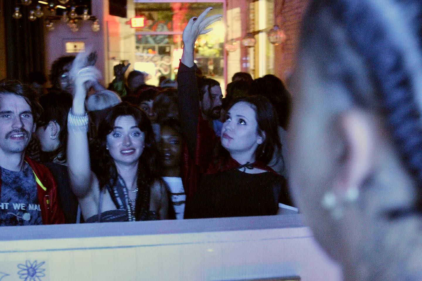 Crowd of people at an event, with a woman in the foreground facing away, and others looking and raising hands.