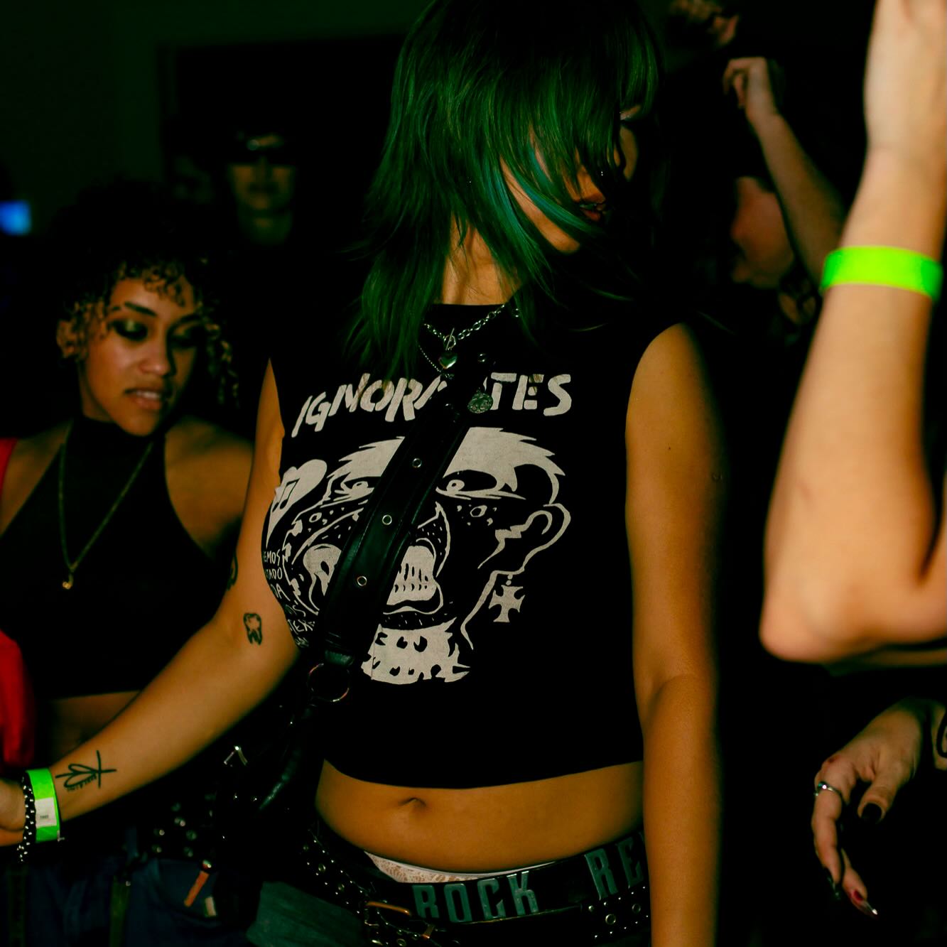 Person with green hair wearing a black sleeveless shirt with a skull graphic, surrounded by others at a dark event.