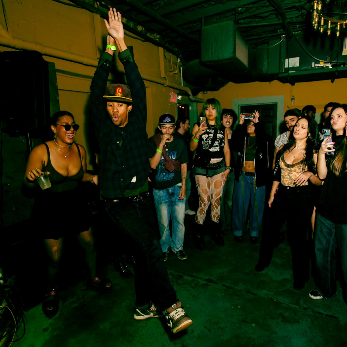 Person dancing with arms raised in a crowded indoor party, others watching and socializing, dim lighting, green ceiling, casual attire.